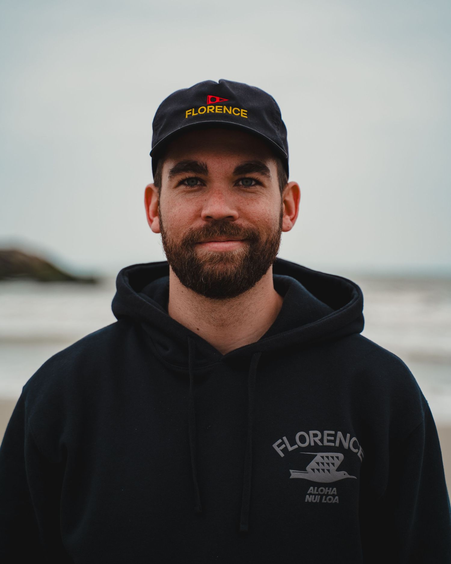 Person wearing a black Florence branded cap and hoodie against a cloudy coastal background.