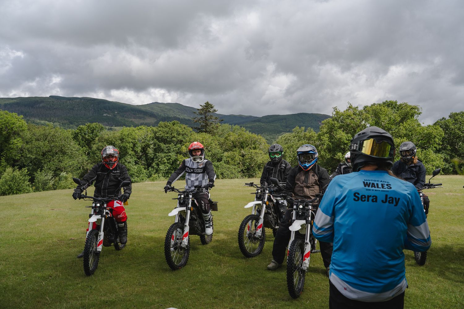 Group of dirt bike riders on a grassy field with helmets and gear, green hills and cloudy sky in background.