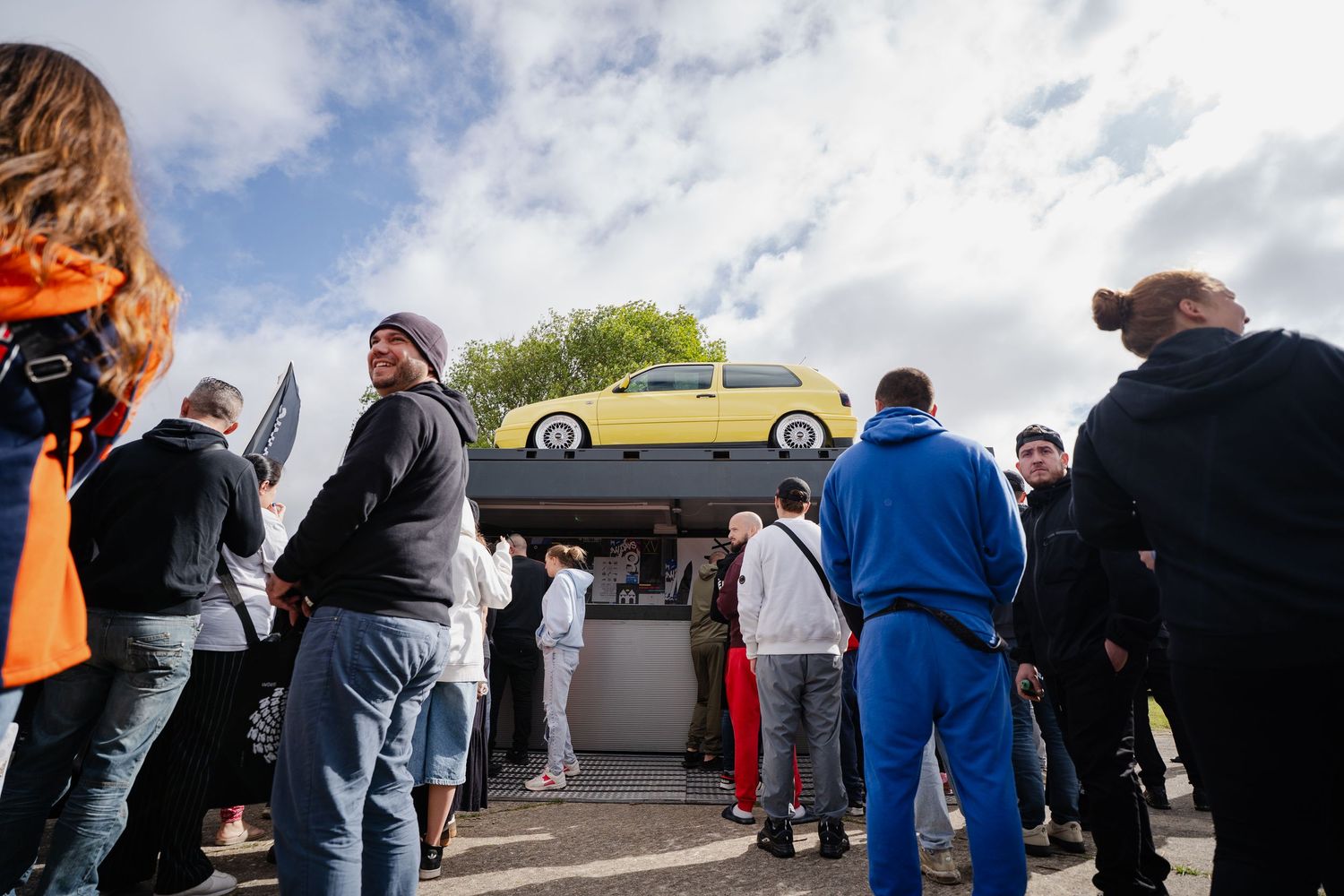 Crowd gathers around a yellow car displayed on an elevated platform at an outdoor car show event.