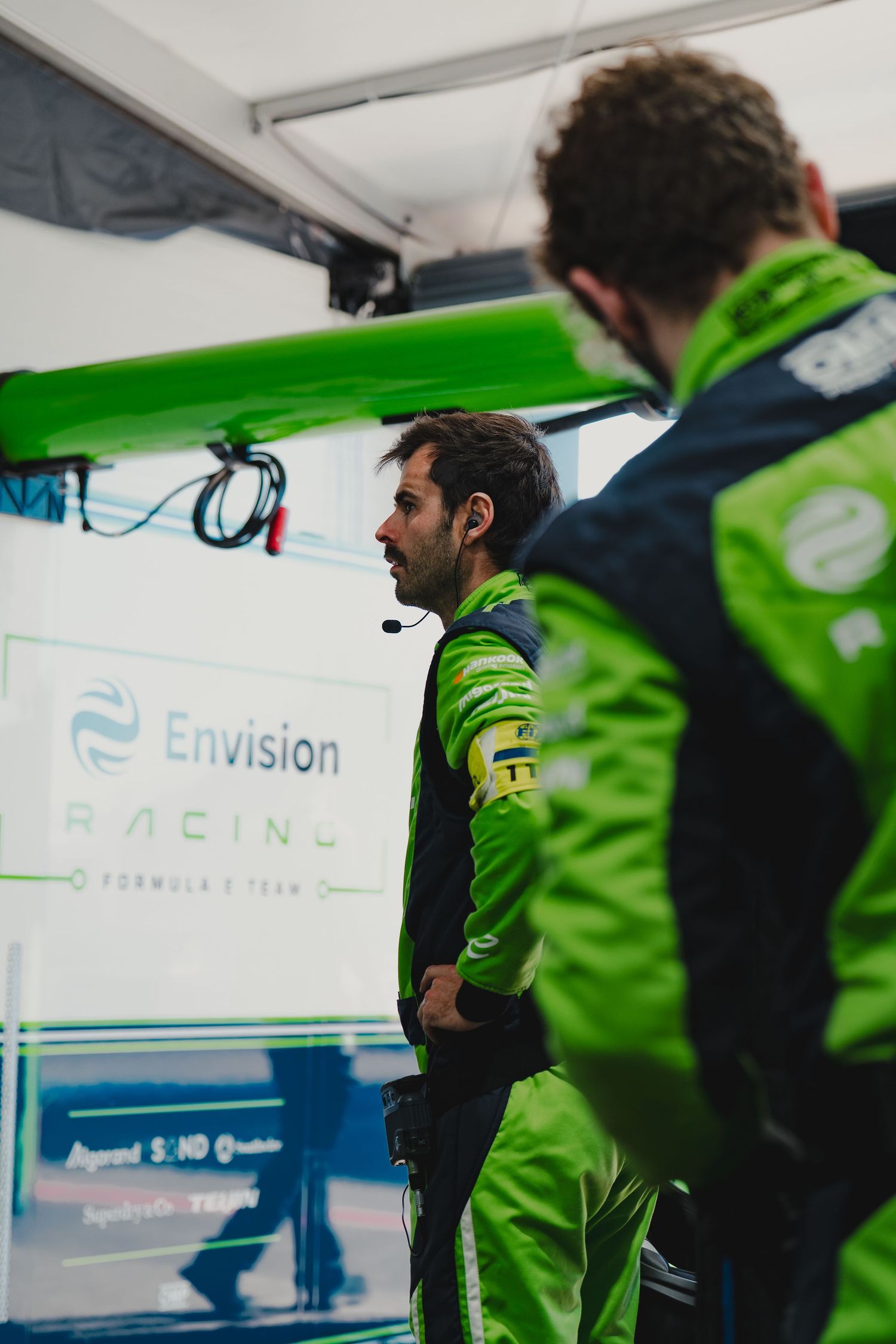 Two Envision Racing Formula E team members in green jackets discuss strategy in the pit garage.