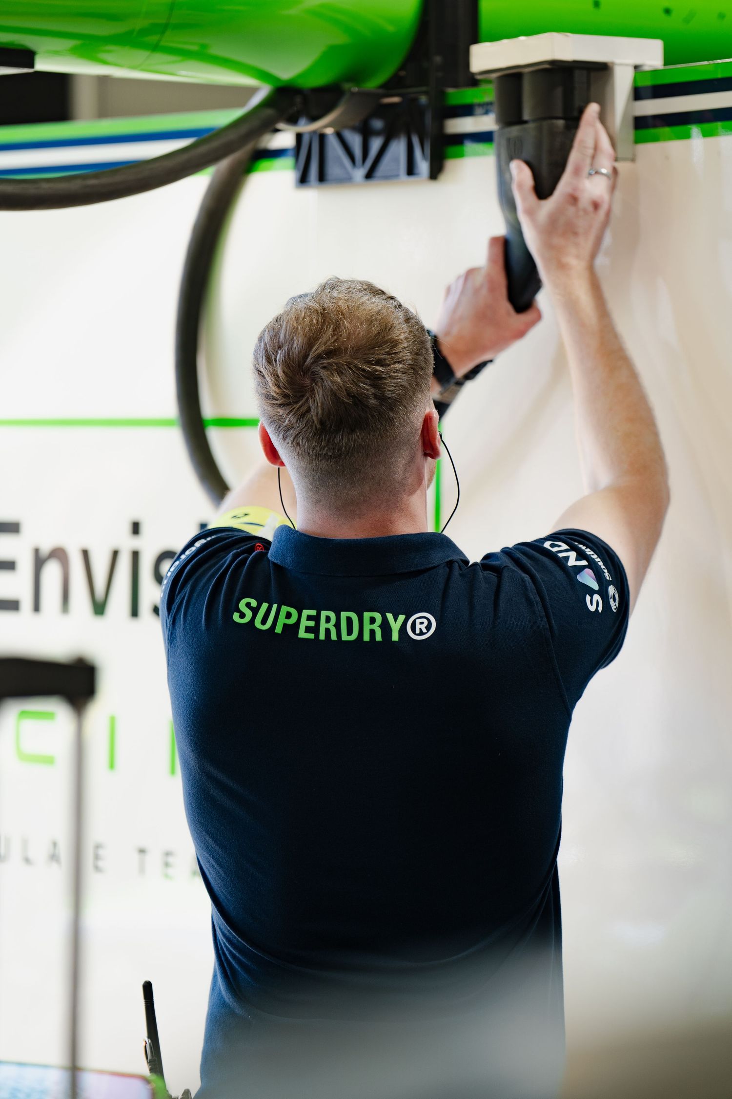A man in a Superdry shirt installs equipment on a wall at an Envision Racing technical facility.