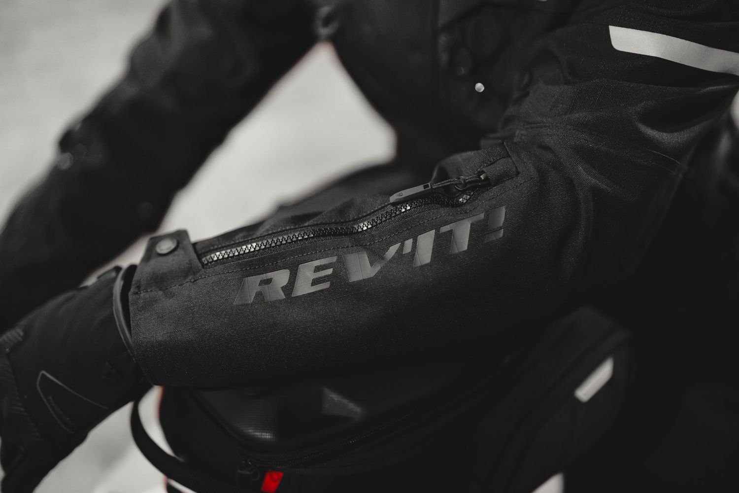 Close-up of a motorcyclist wearing a black REV'IT! armored riding jacket with zippered pockets.