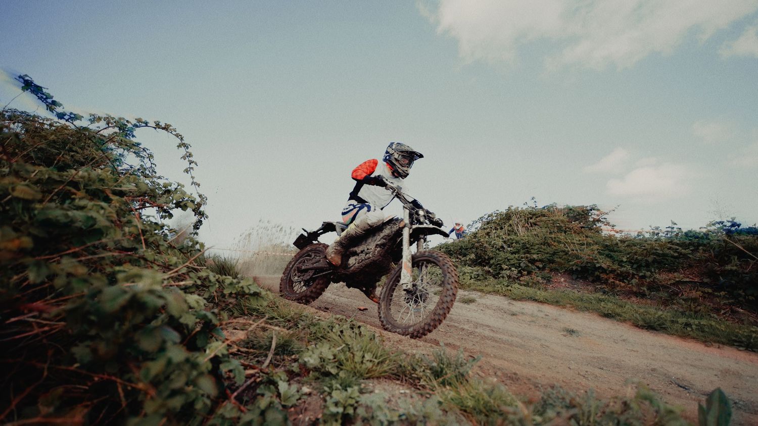 A motocross rider on a muddy dirt bike navigates a dusty off-road trail surrounded by green shrubs under a cloudy sky.