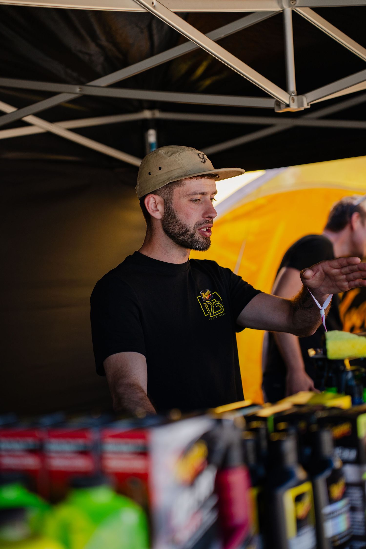 Man in black t-shirt and tan cap working at an outdoor vendor booth under a canopy tent.