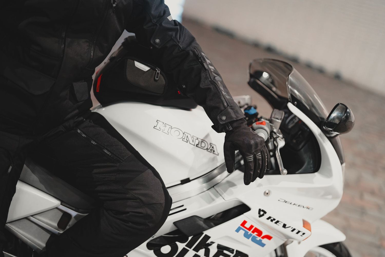 Motorcyclist in black riding gear seated on a white Honda sport motorcycle on a cobblestone surface.