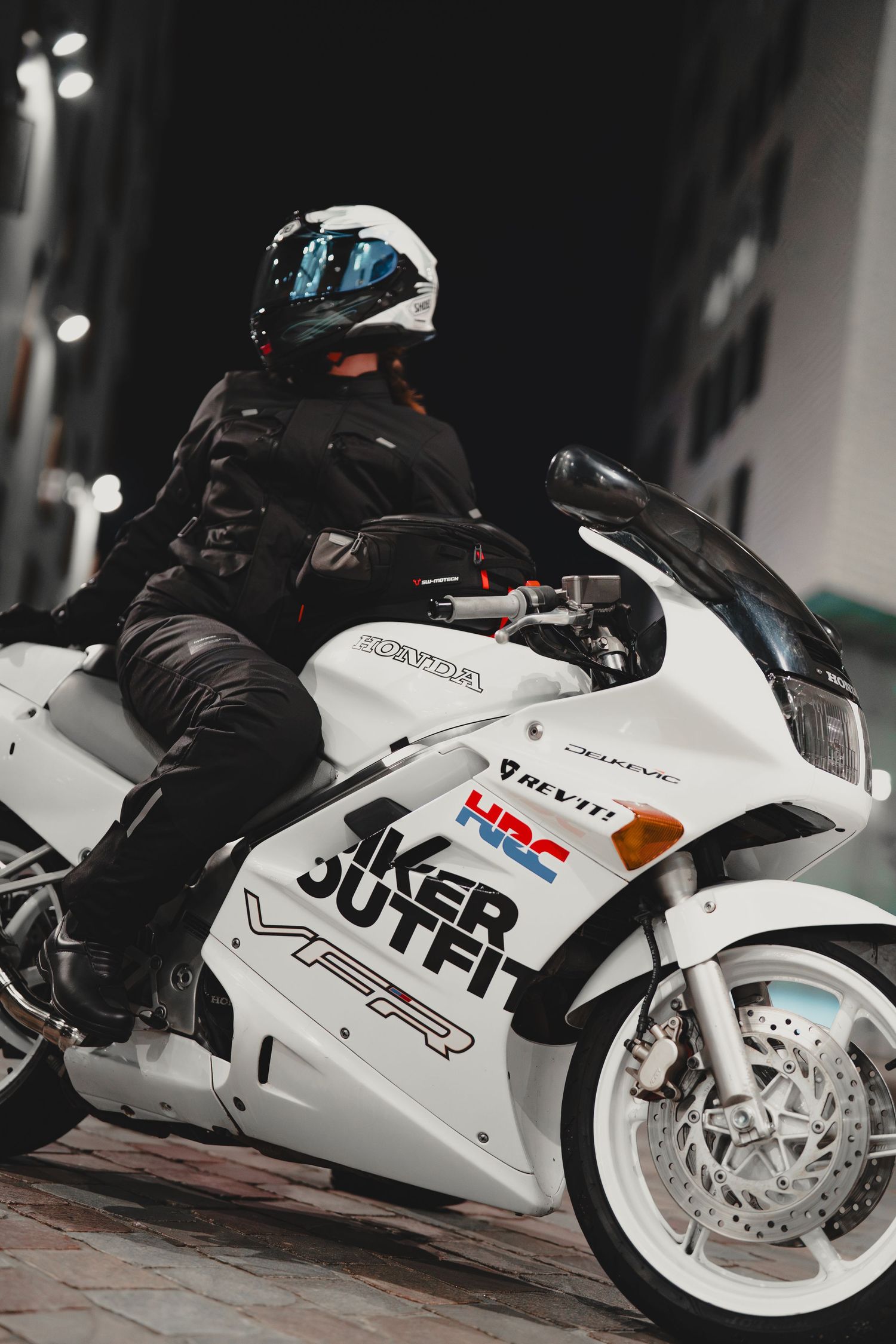 A motorcyclist in black leather gear and white helmet rides a white Honda VFR sport bike at night.