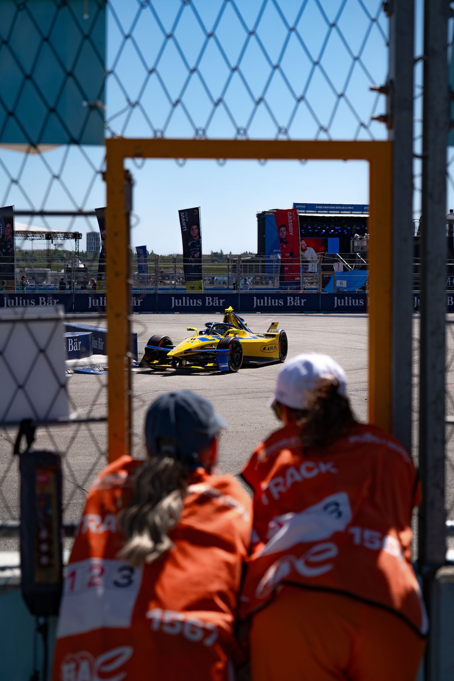 Formula E yellow race car speeds past track marshals in orange uniforms at Julius Baer sponsored street circuit.
