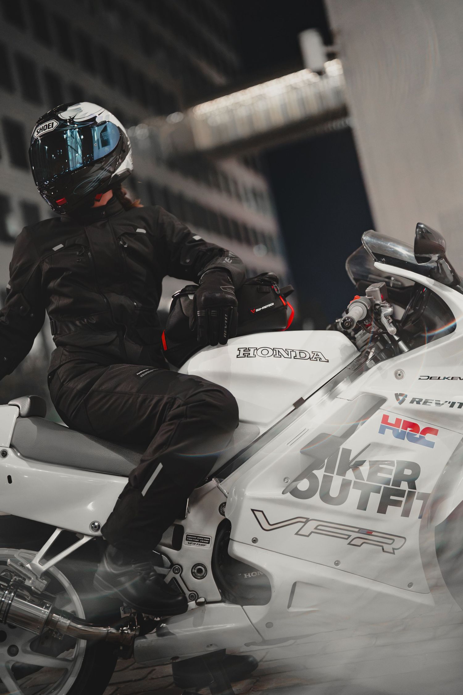 Motorcyclist in black gear and helmet riding a white Honda VFR sport bike with HRC and Biker Outfit branding.