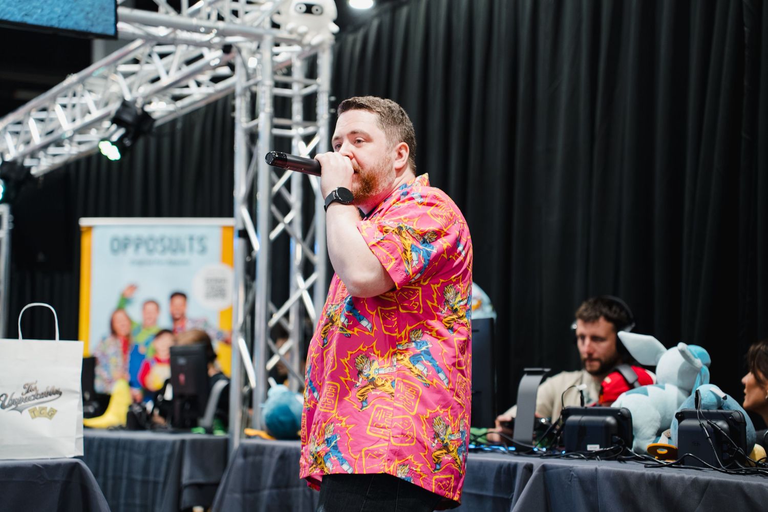 Man in colorful floral shirt performing on stage with microphone at outdoor event with DJ in background.