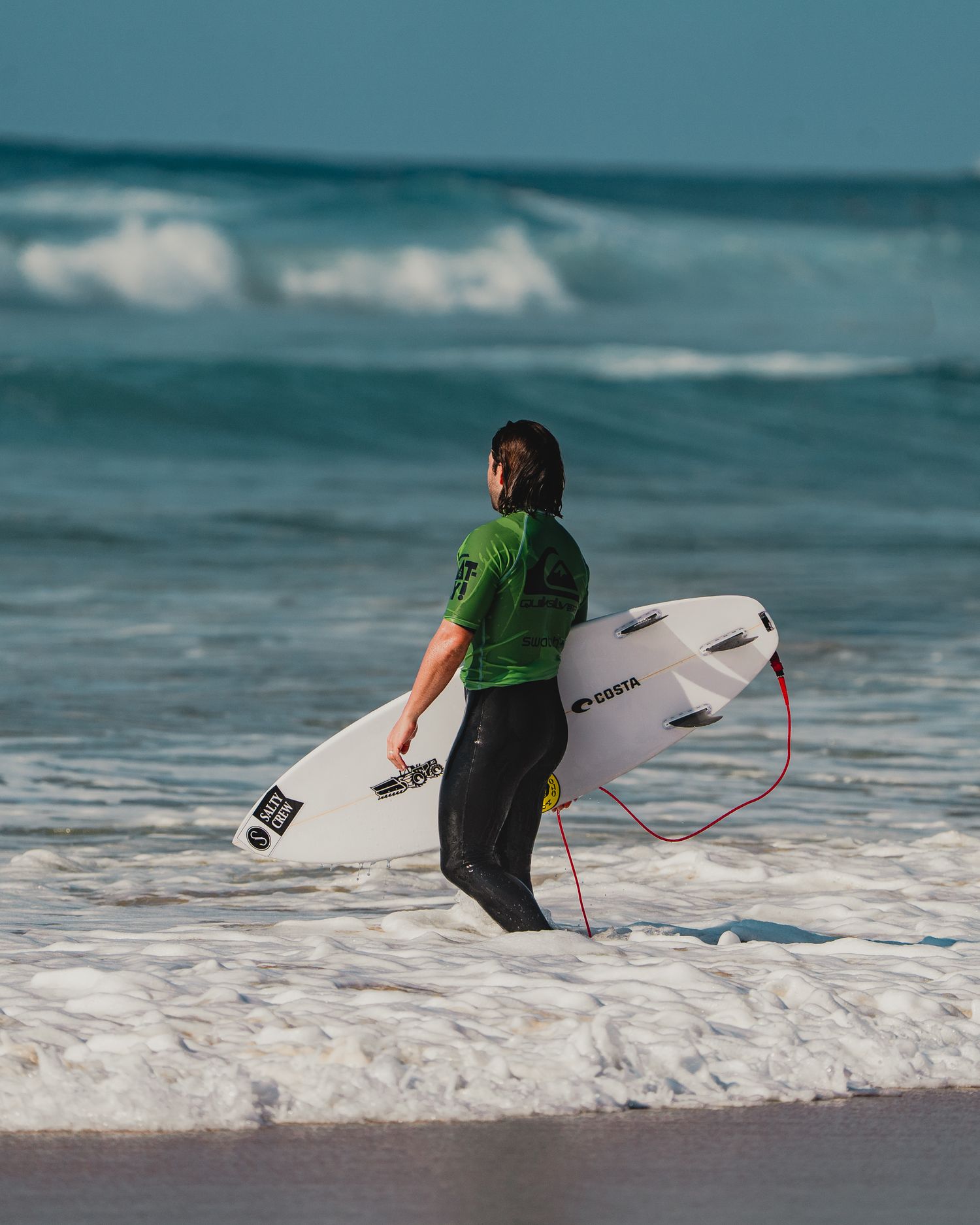 A surfer in a wetsuit runs into the ocean waves with their surfboard.