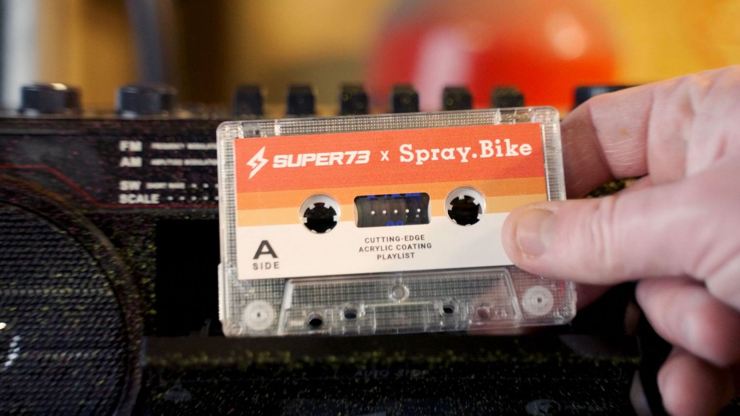 Super73 x Spray.Bike branded cassette tape resting on a vintage boombox, held by a hand.
