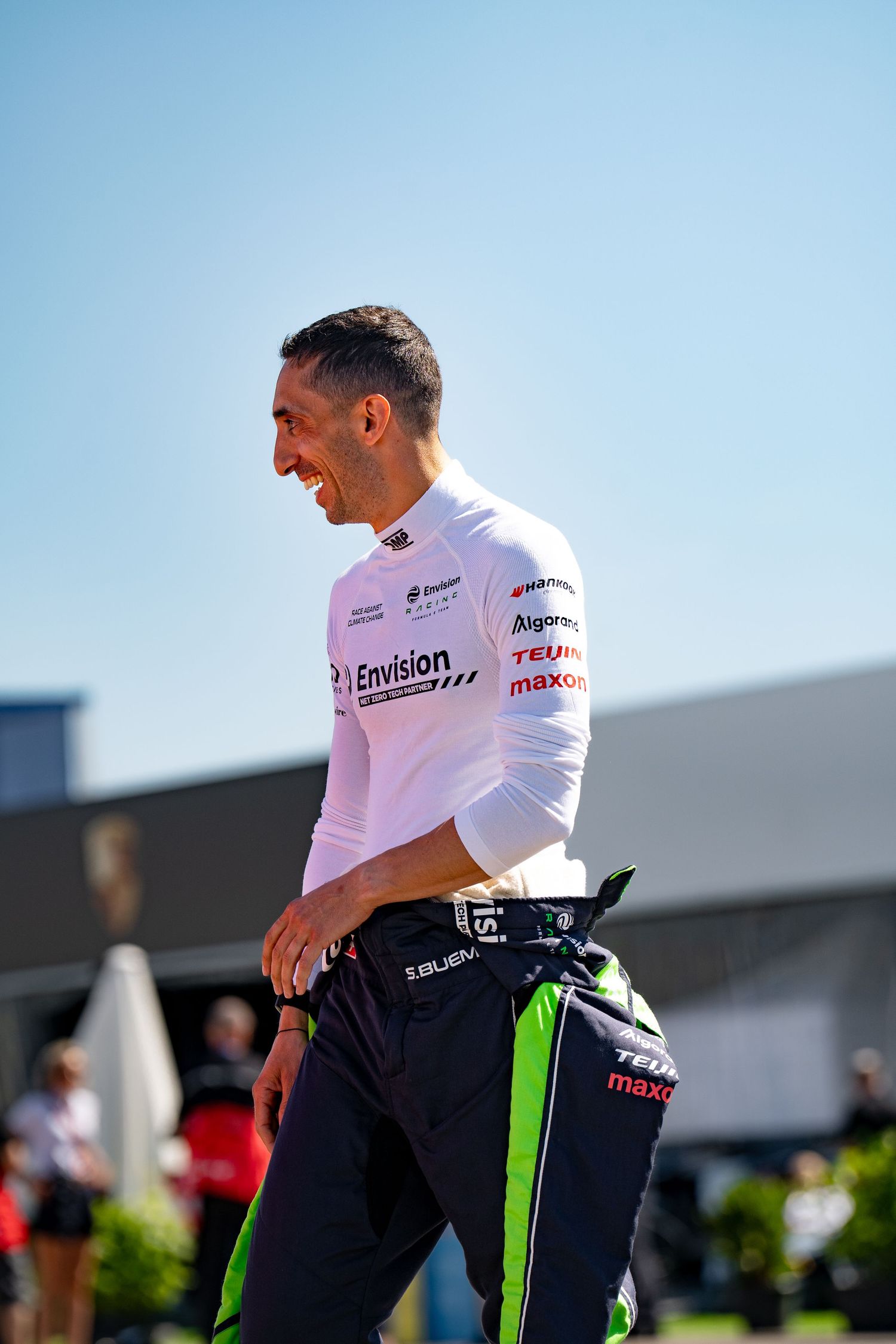 Racing driver in Envision team uniform smiling outdoors at a motorsport event on a sunny day.