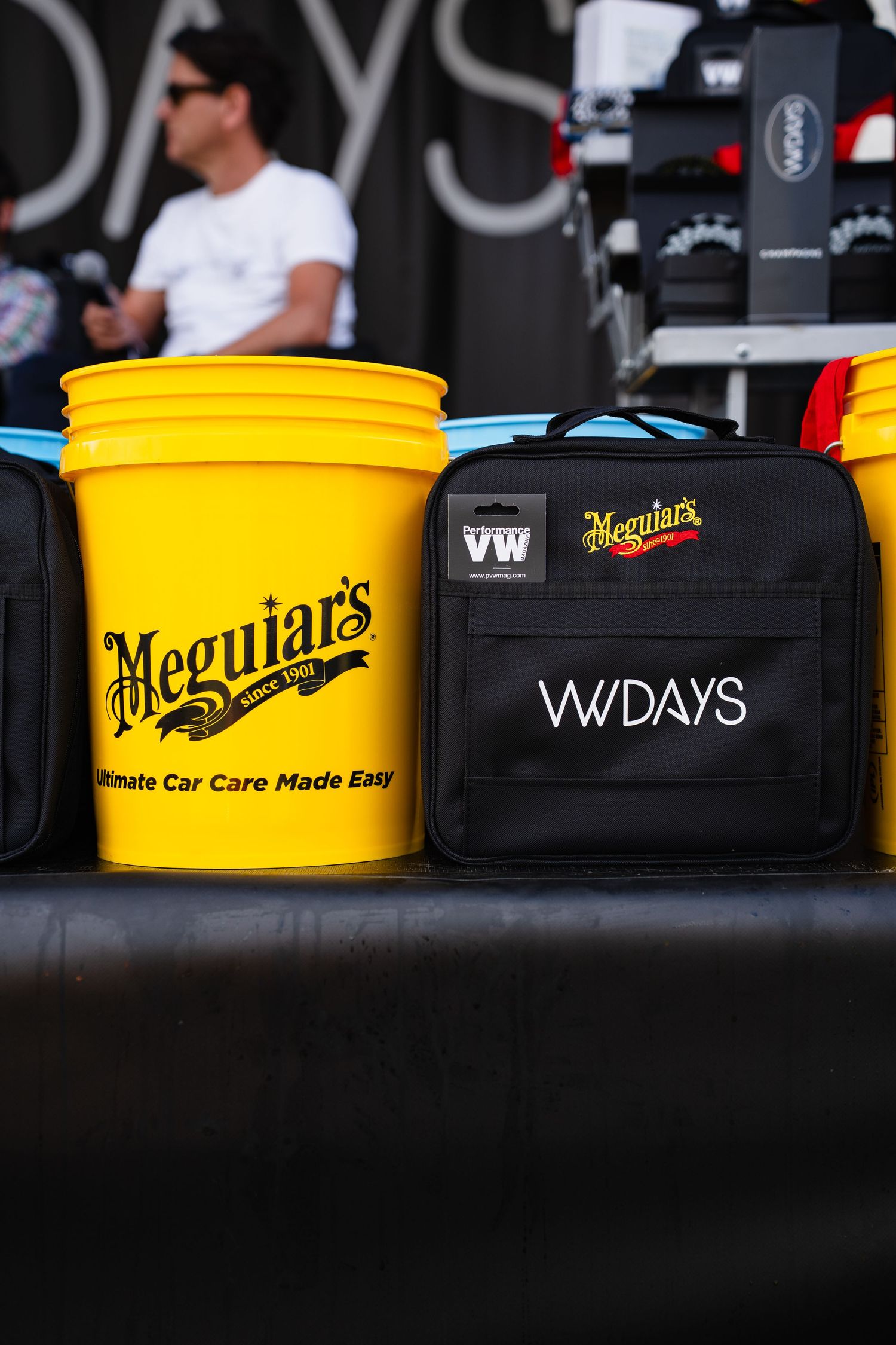 Meguiar's yellow car care bucket and branded VW Days bag displayed on a table at an automotive detailing event.