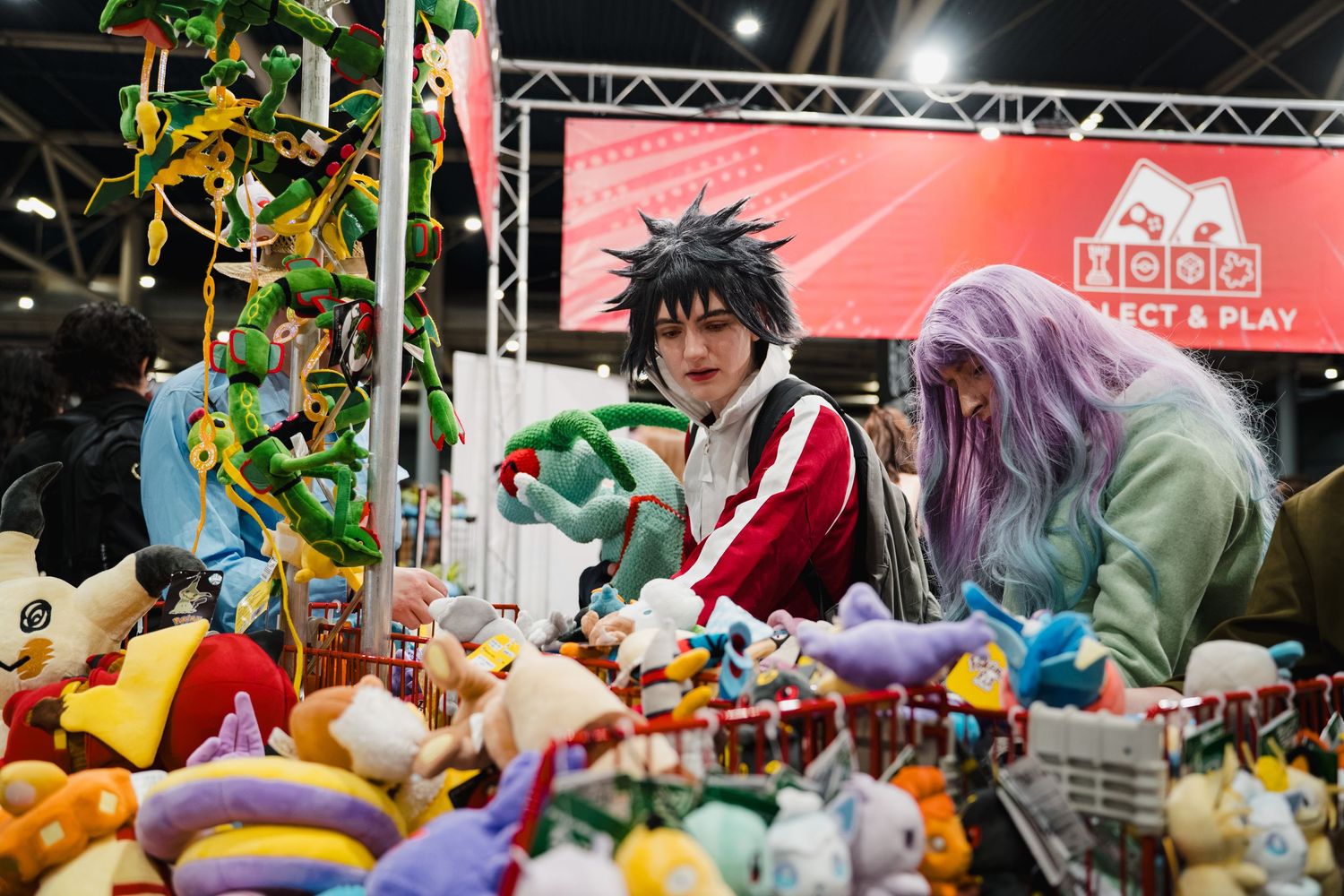 Two cosplayers browse colorful plush toys at a gaming convention booth with a red banner backdrop.