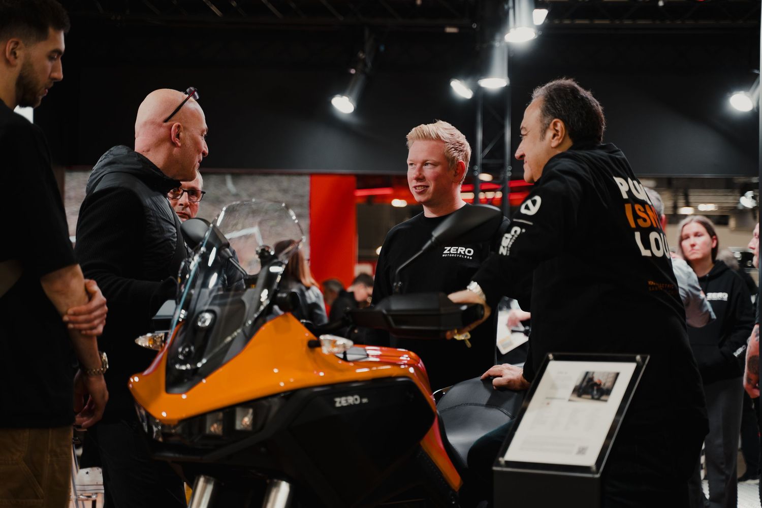 Indoor motorcycle show with orange and black bikes on display under bright showroom lighting.