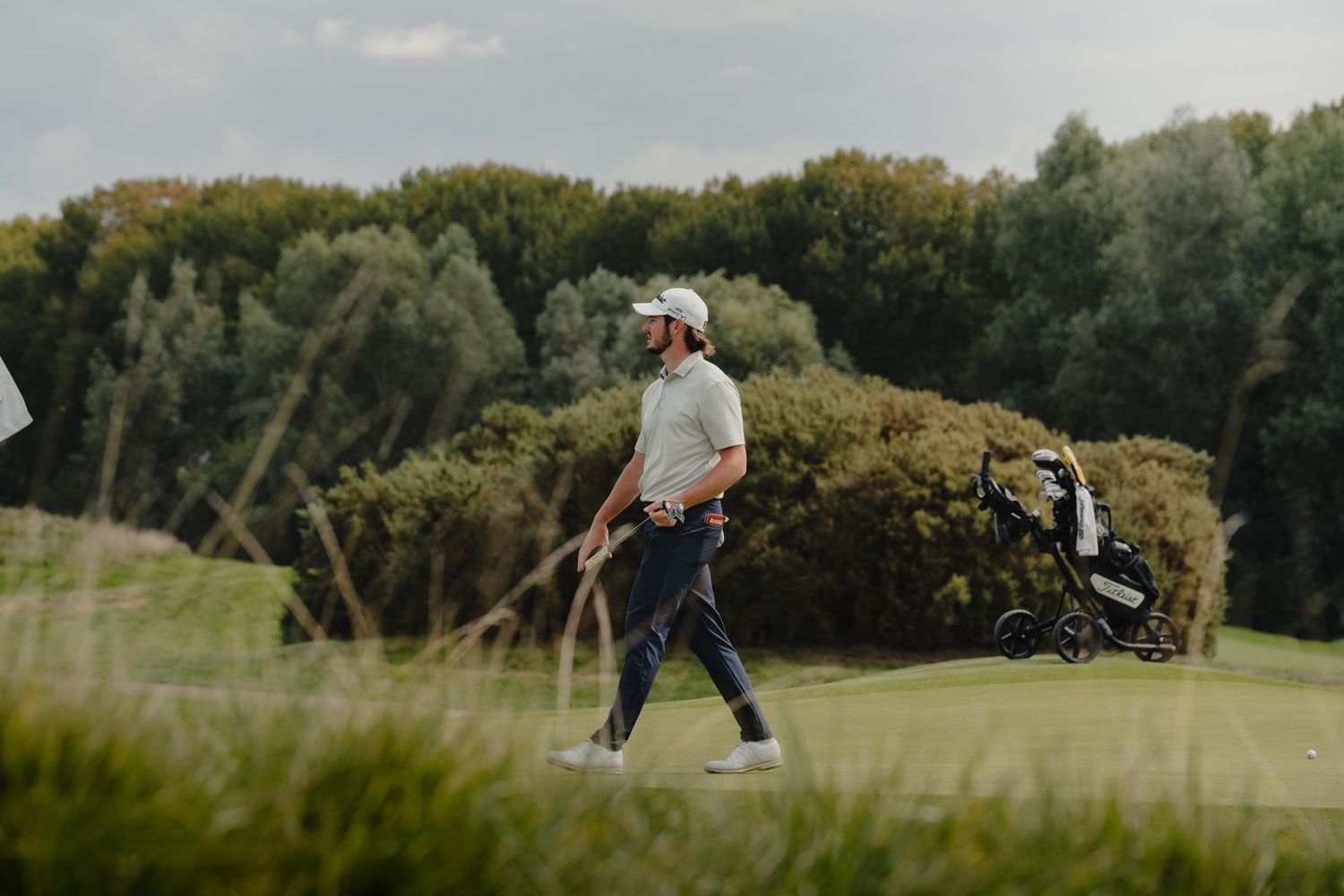 Golfer walking on lush green course with golf bag and scenic tree backdrop.