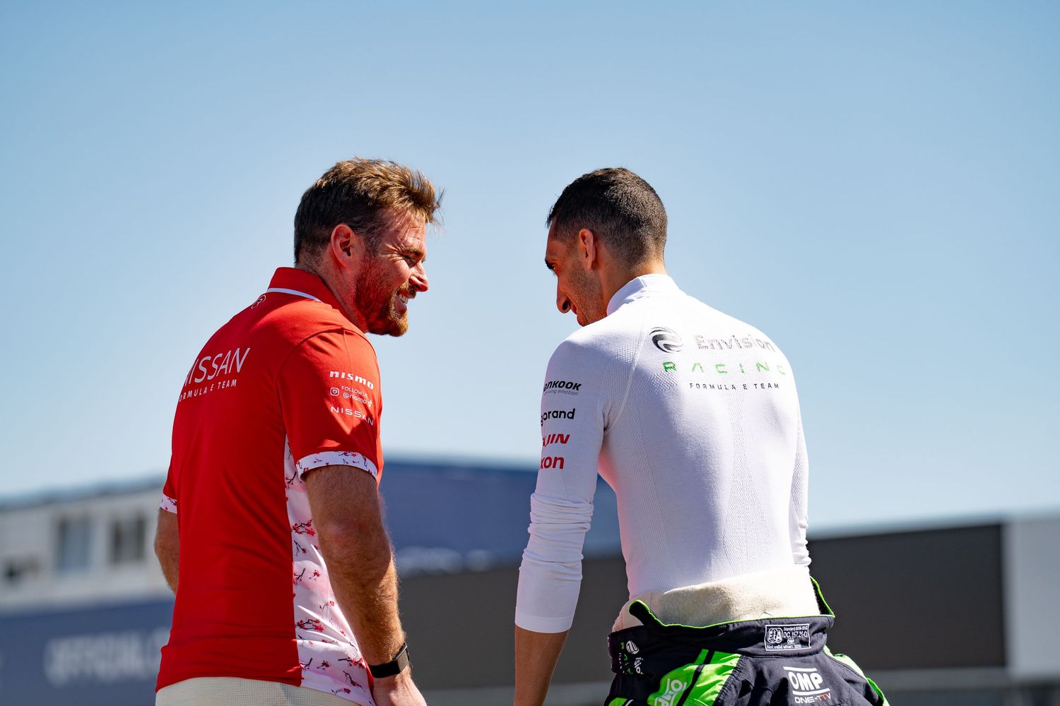 Two Formula E drivers in team gear having a conversation outdoors at a racing circuit on a sunny day.