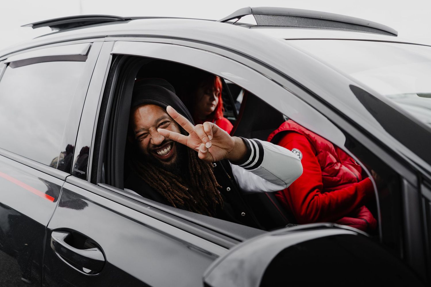 Man with dreadlocks in red jacket flashing peace sign from black car window, smiling cheerfully.