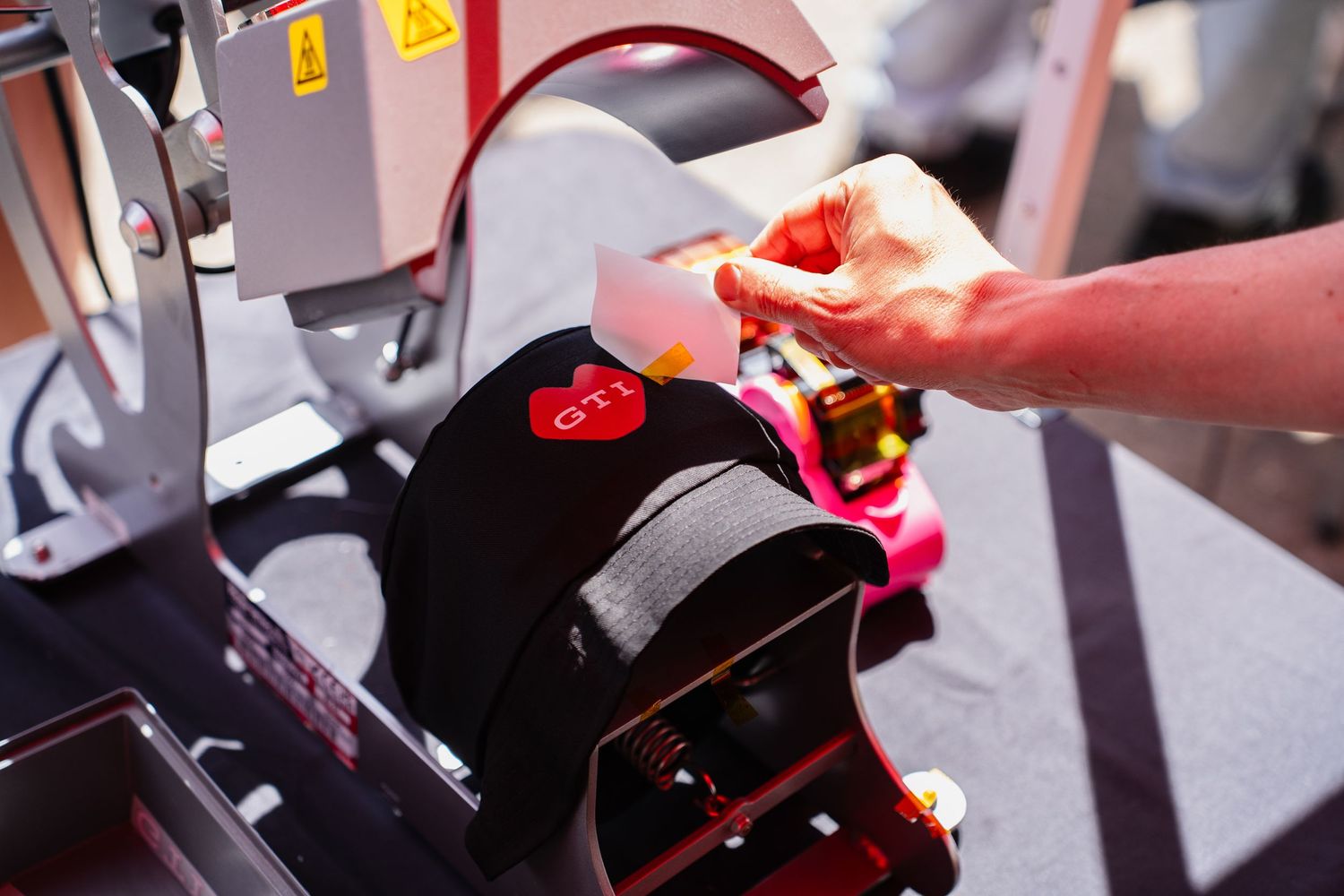 Hands applying a GTI logo patch to a black cap using a heat press machine.