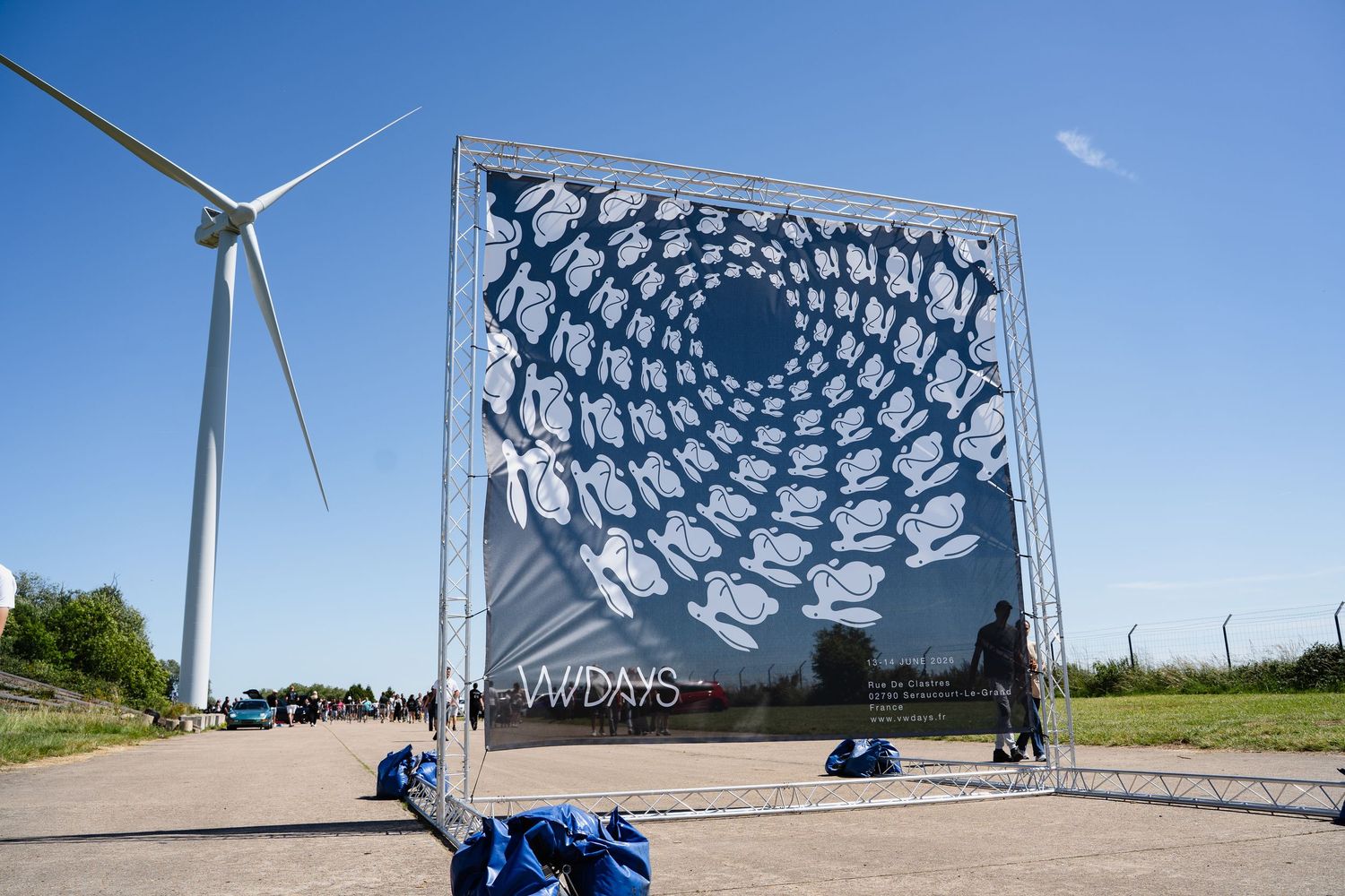 Large mirrored art installation with wind turbine against blue sky at outdoor festival.