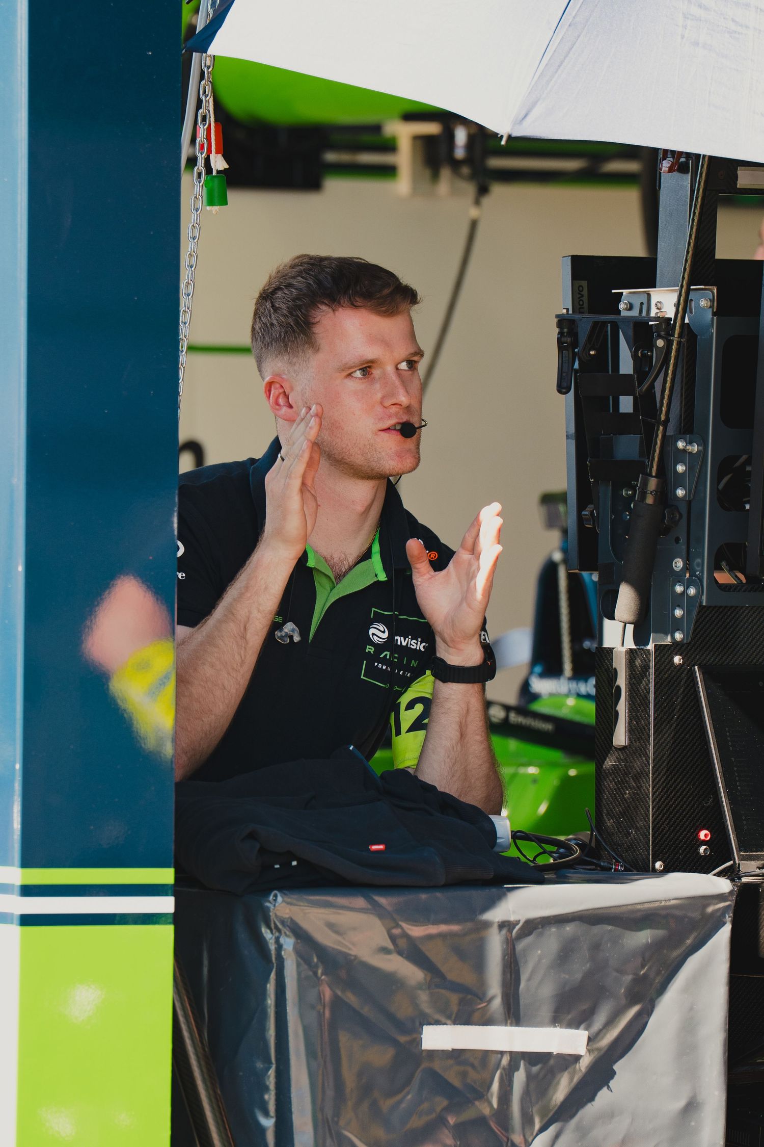 Racing team engineer gestures animatedly at monitors in a motorsport pit garage, wearing a green and black team uniform.