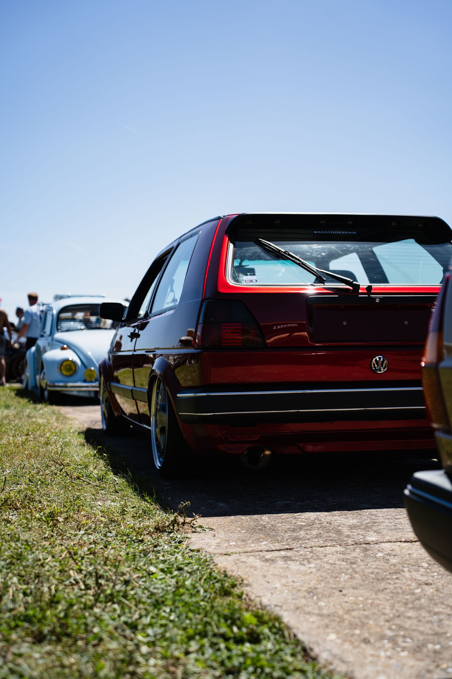 Red VW Golf mk2 lowered stance at a car show with a classic VW Beetle visible in the background.