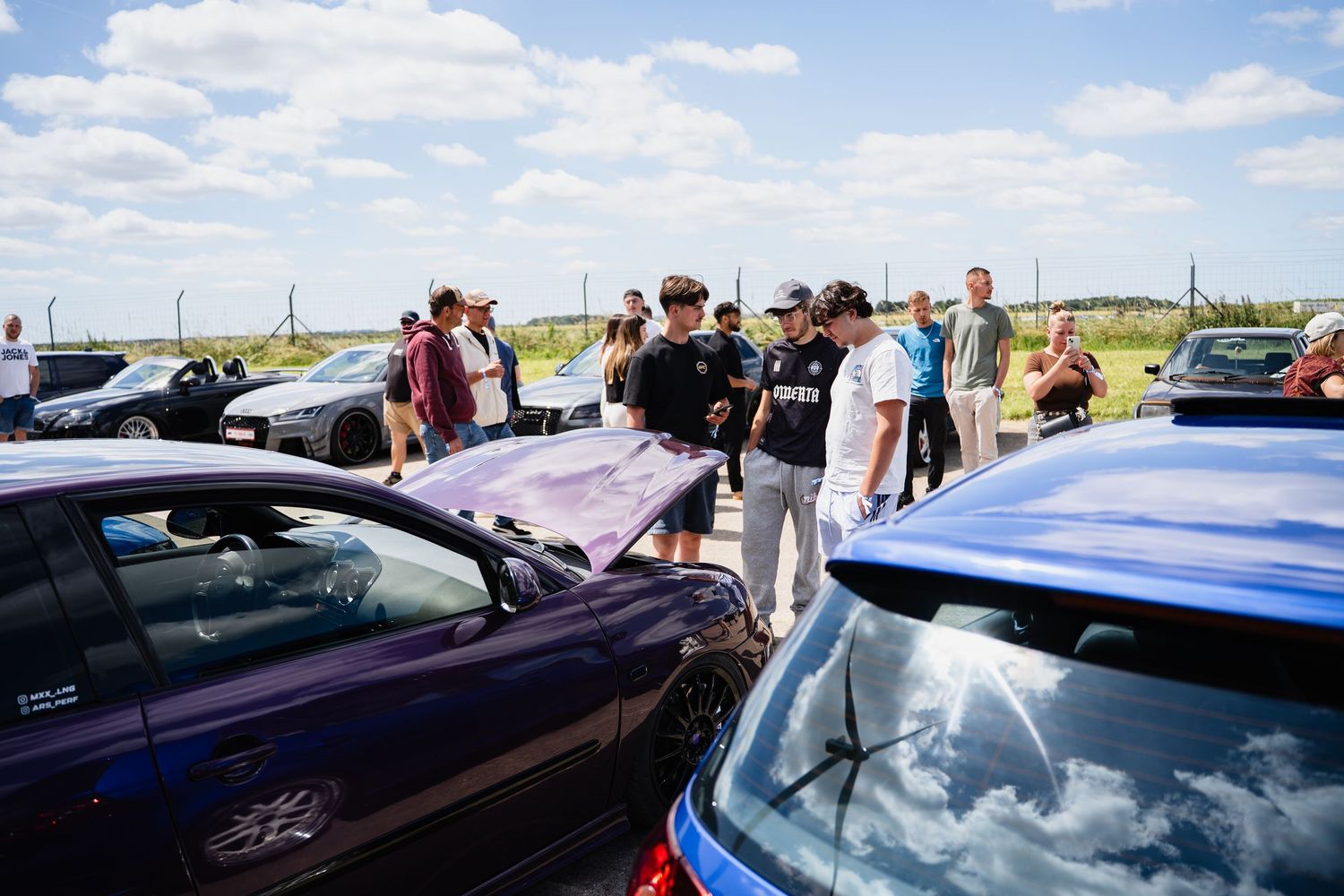 Car enthusiasts gather outdoors at an automotive meet, examining vehicles on a sunny day with blue skies.