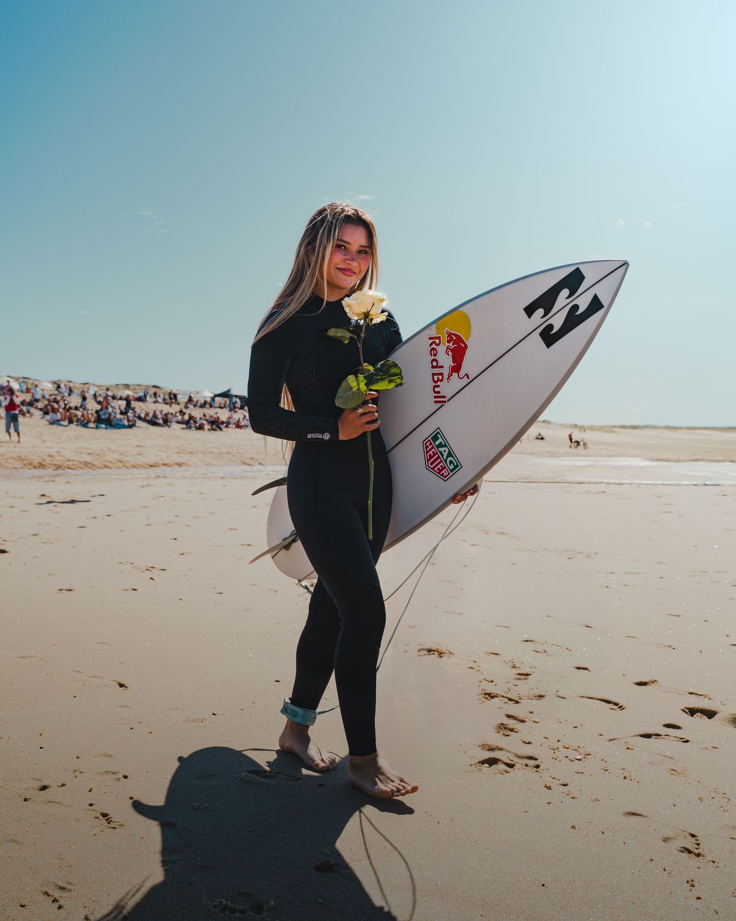 A surfer carrying a white surfboard walks along a sandy beach on a sunny day.