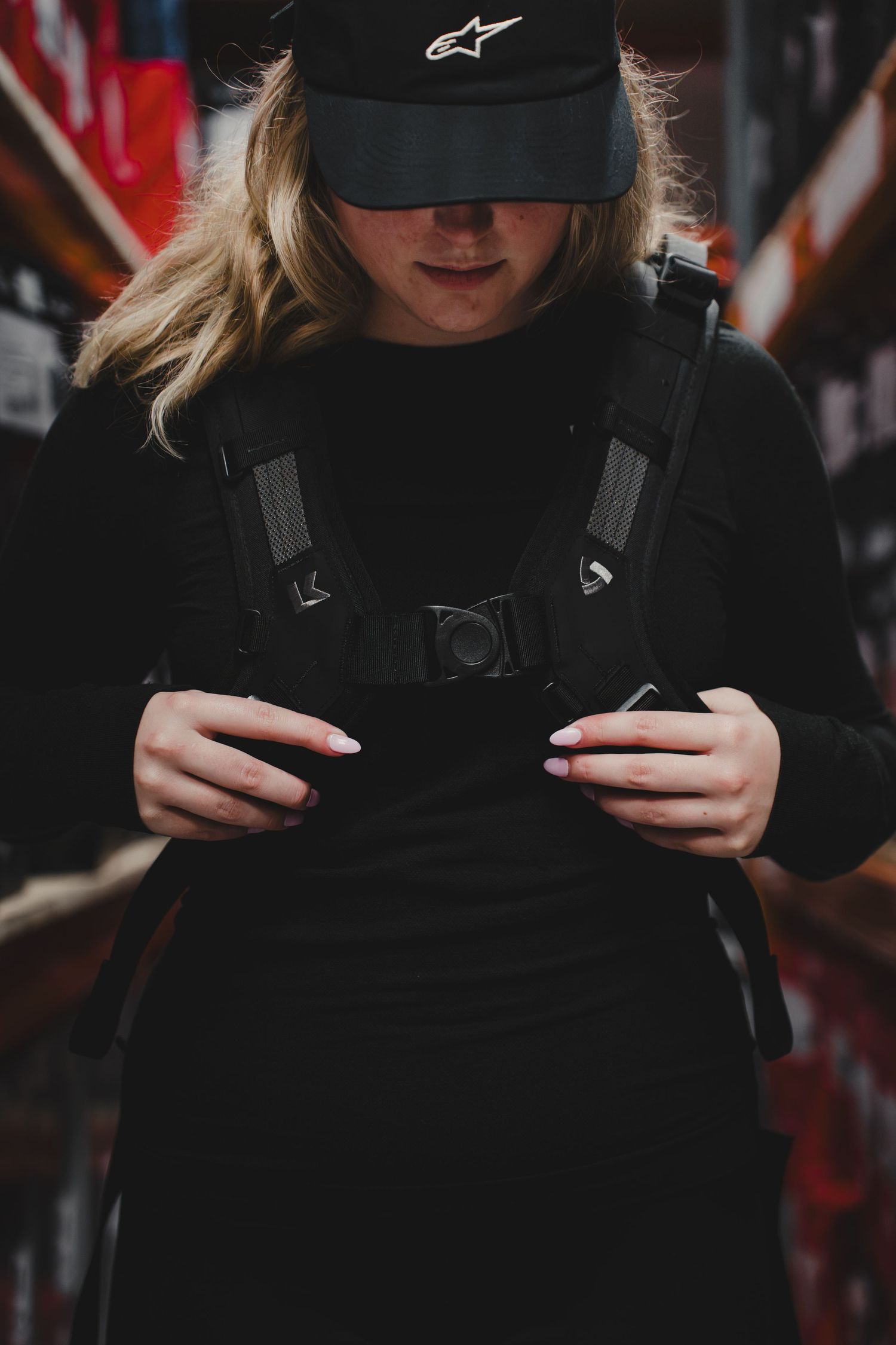 Young woman in black outfit and Alpinestars cap adjusting backpack straps indoors.