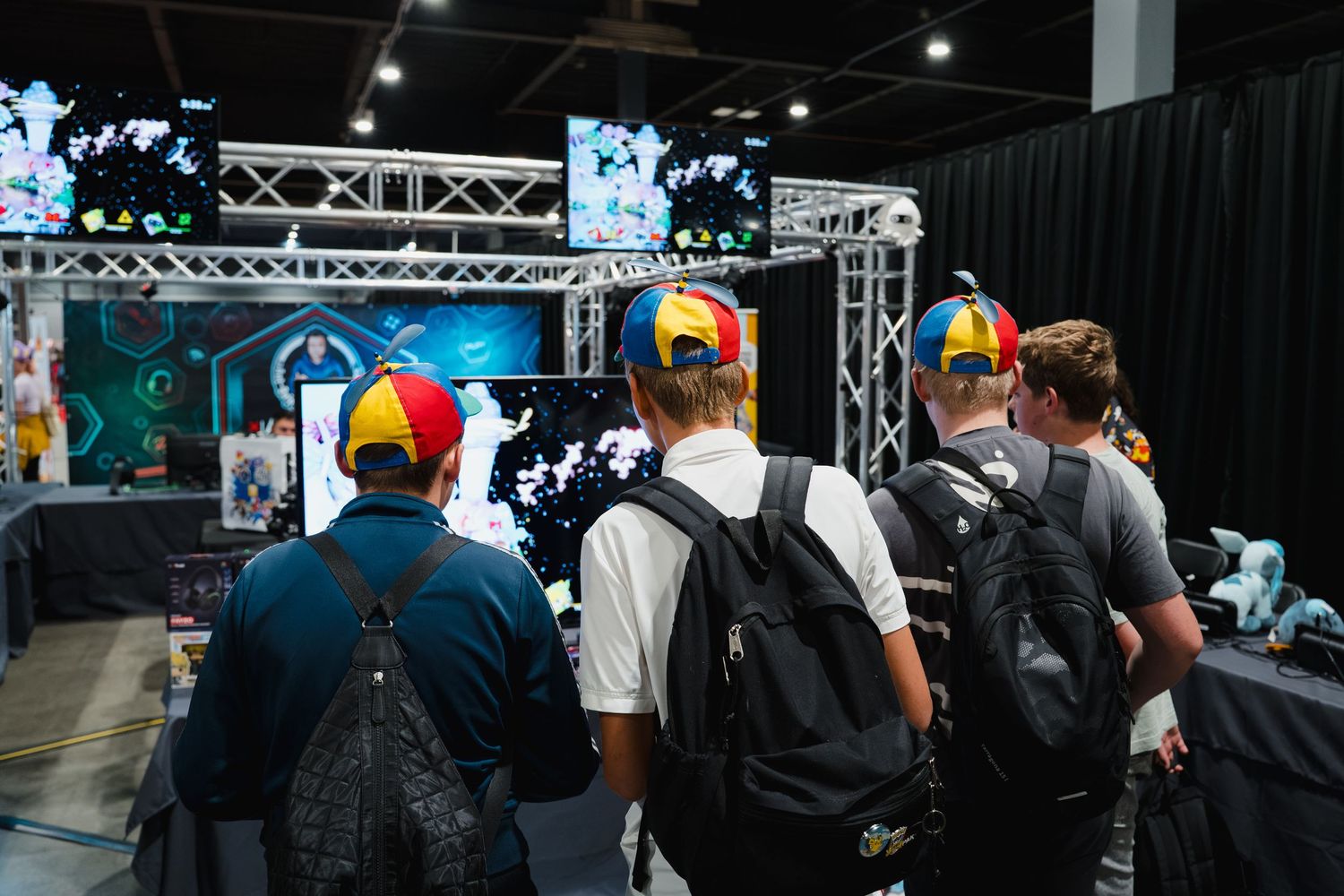 Group of young people wearing colorful caps viewing gaming displays at an indoor convention event.