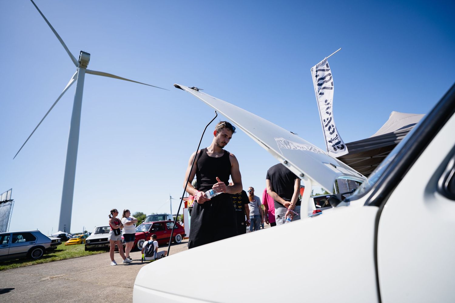 Person examining a vintage white car at an outdoor event with a wind turbine in the background on a sunny day.