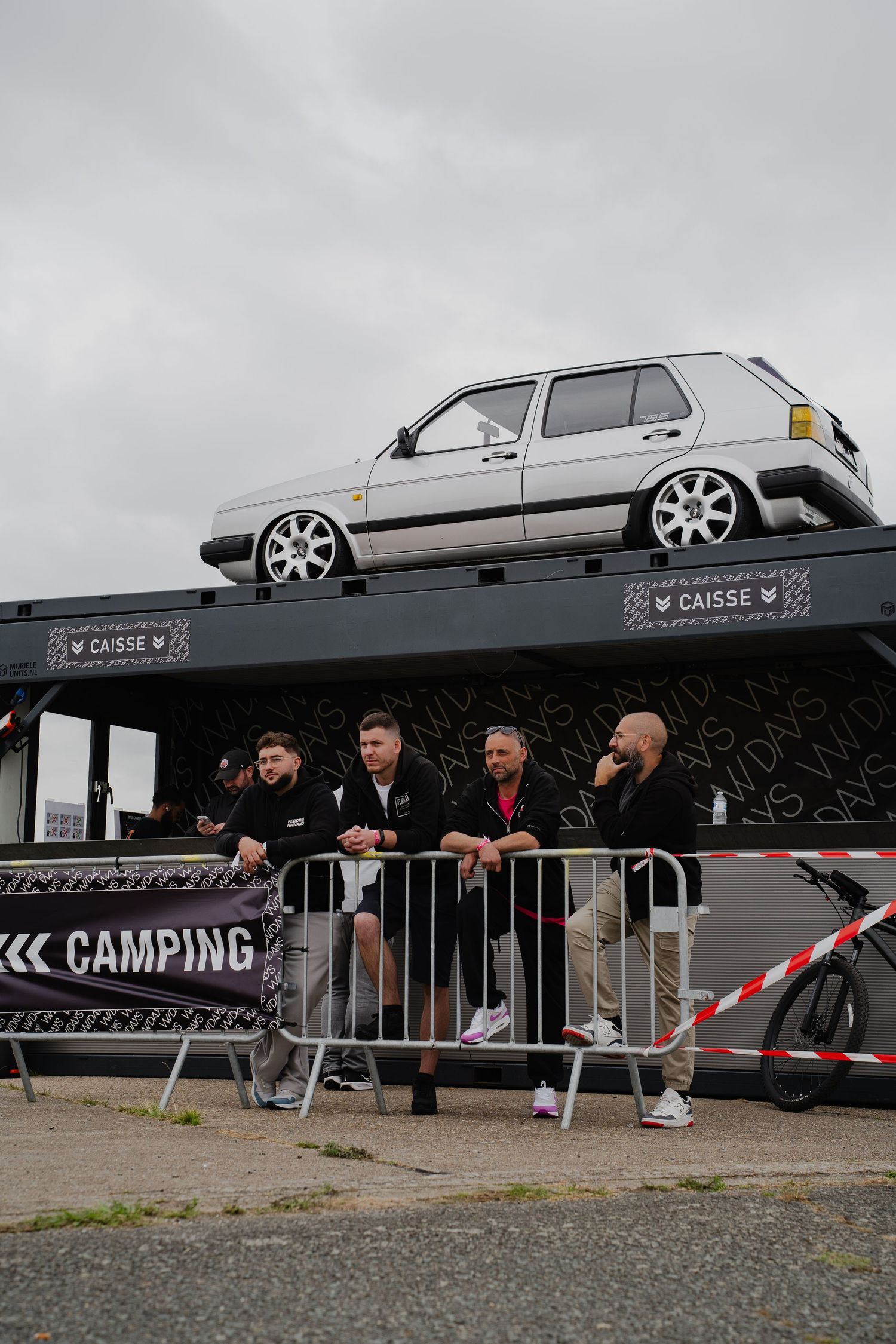 Four men stand near a barrier at a camping event, with a white VW Golf displayed on an elevated platform above them.