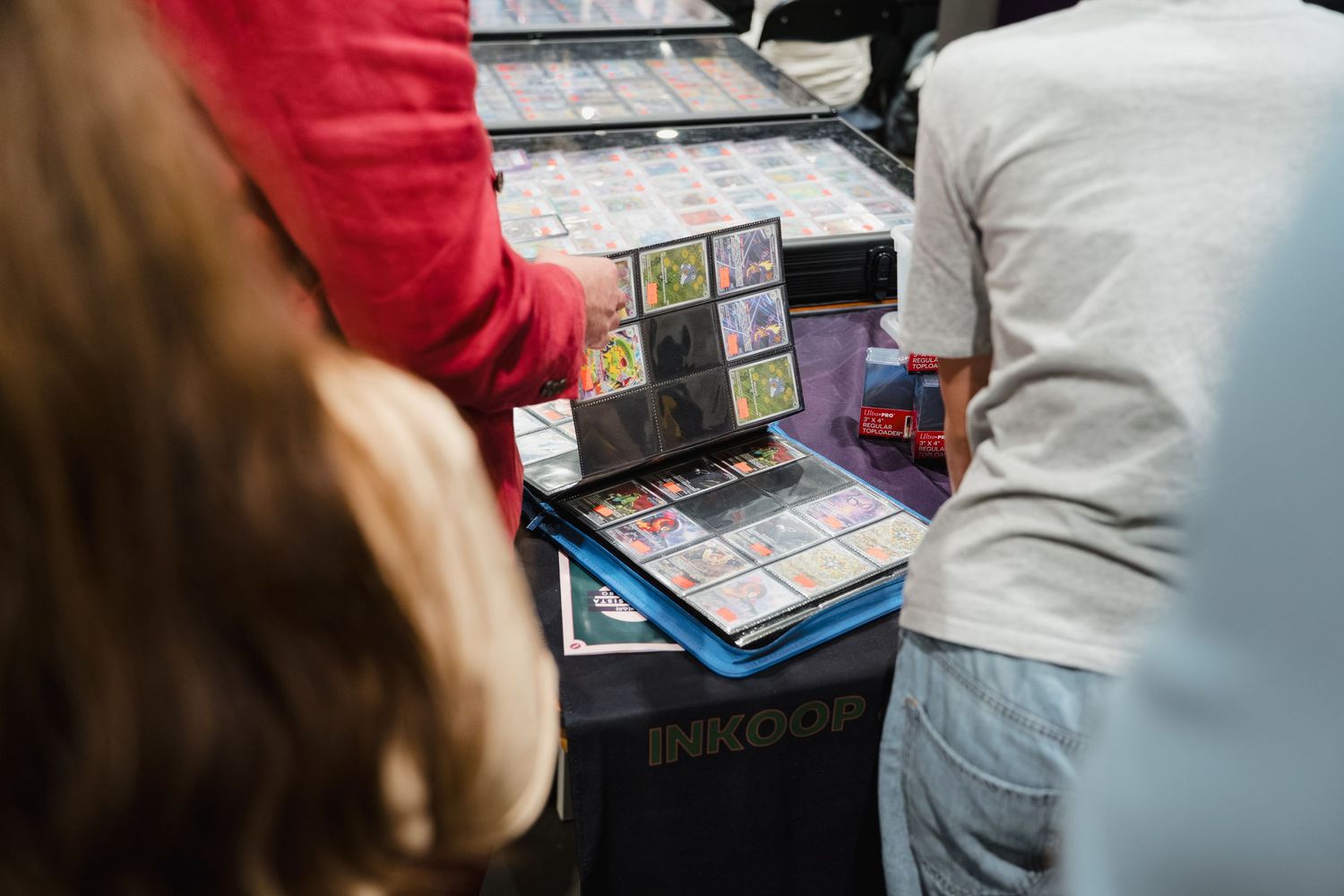 People browsing trading card binders at an Inkoop vendor table at a convention or card show.
