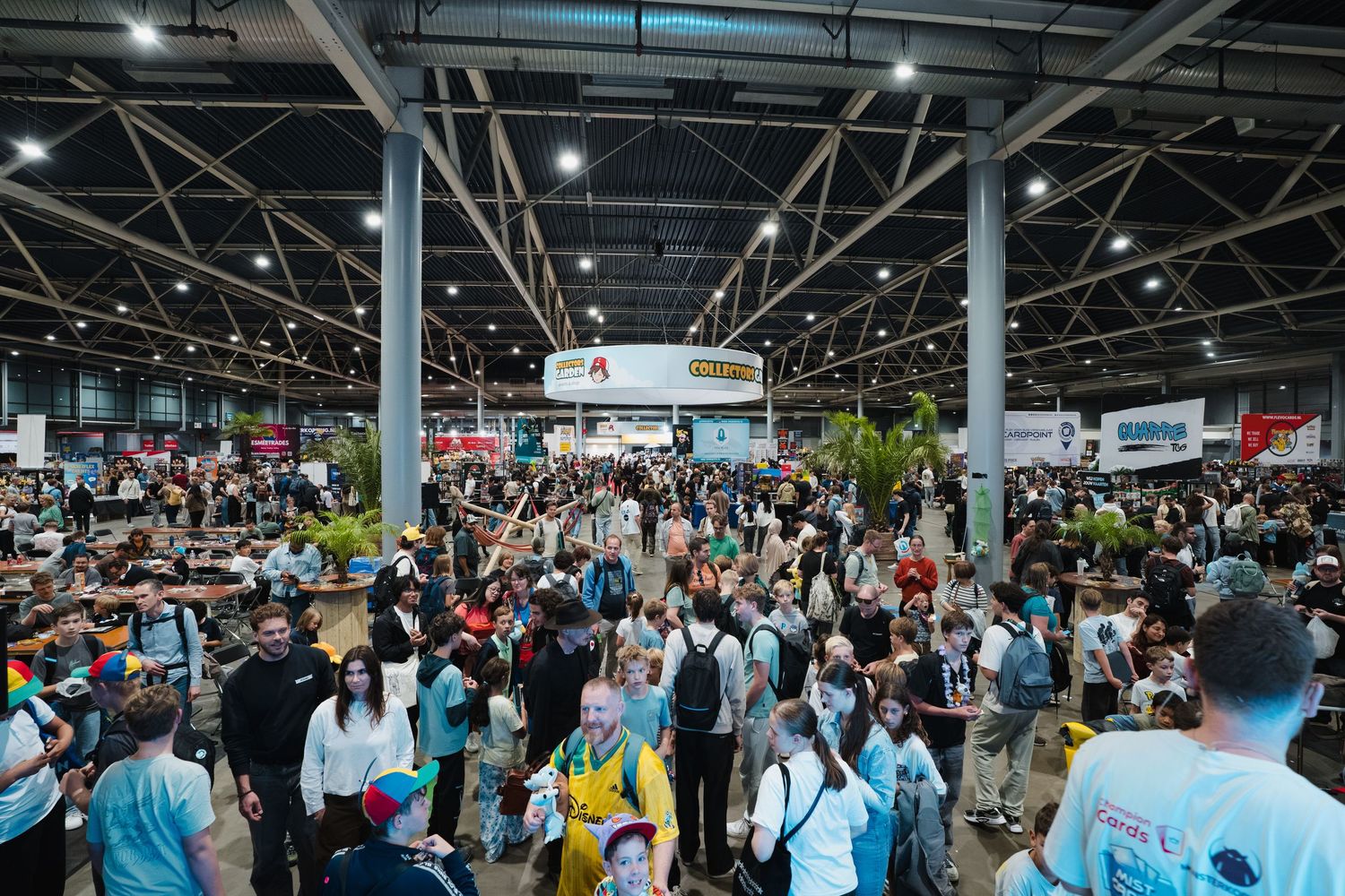 Crowded convention hall filled with attendees browsing exhibits under industrial ceiling lights.