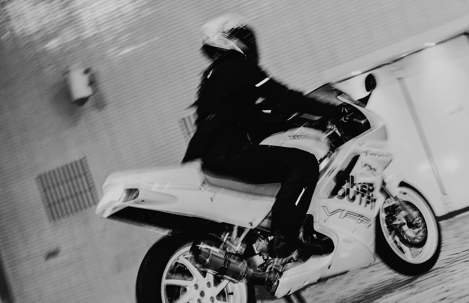 Black and white photo of a motorcyclist in helmet and jacket riding a white sport motorcycle on a road.