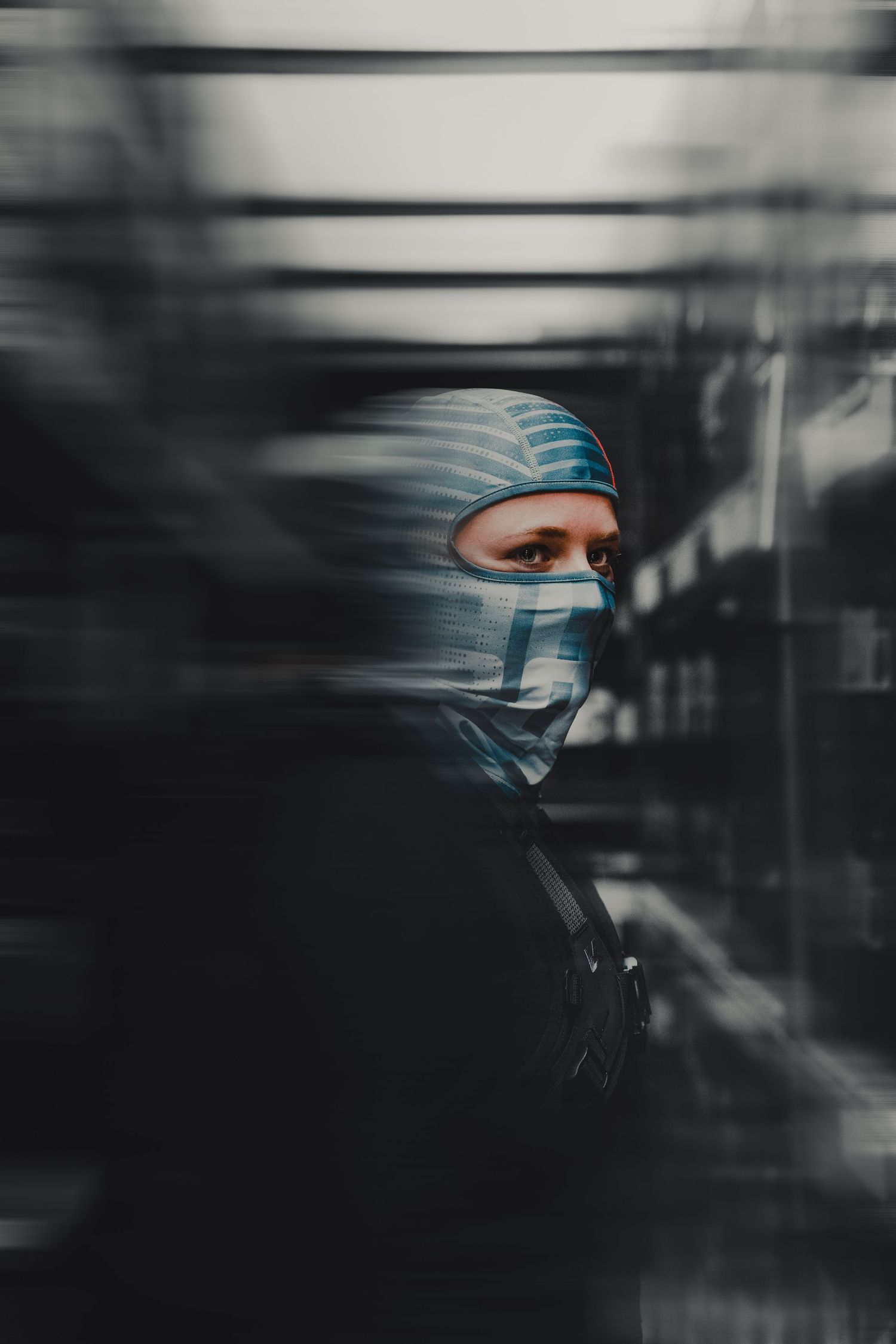 Person wearing blue striped face mask and matching head covering, photographed through blurred foreground elements.