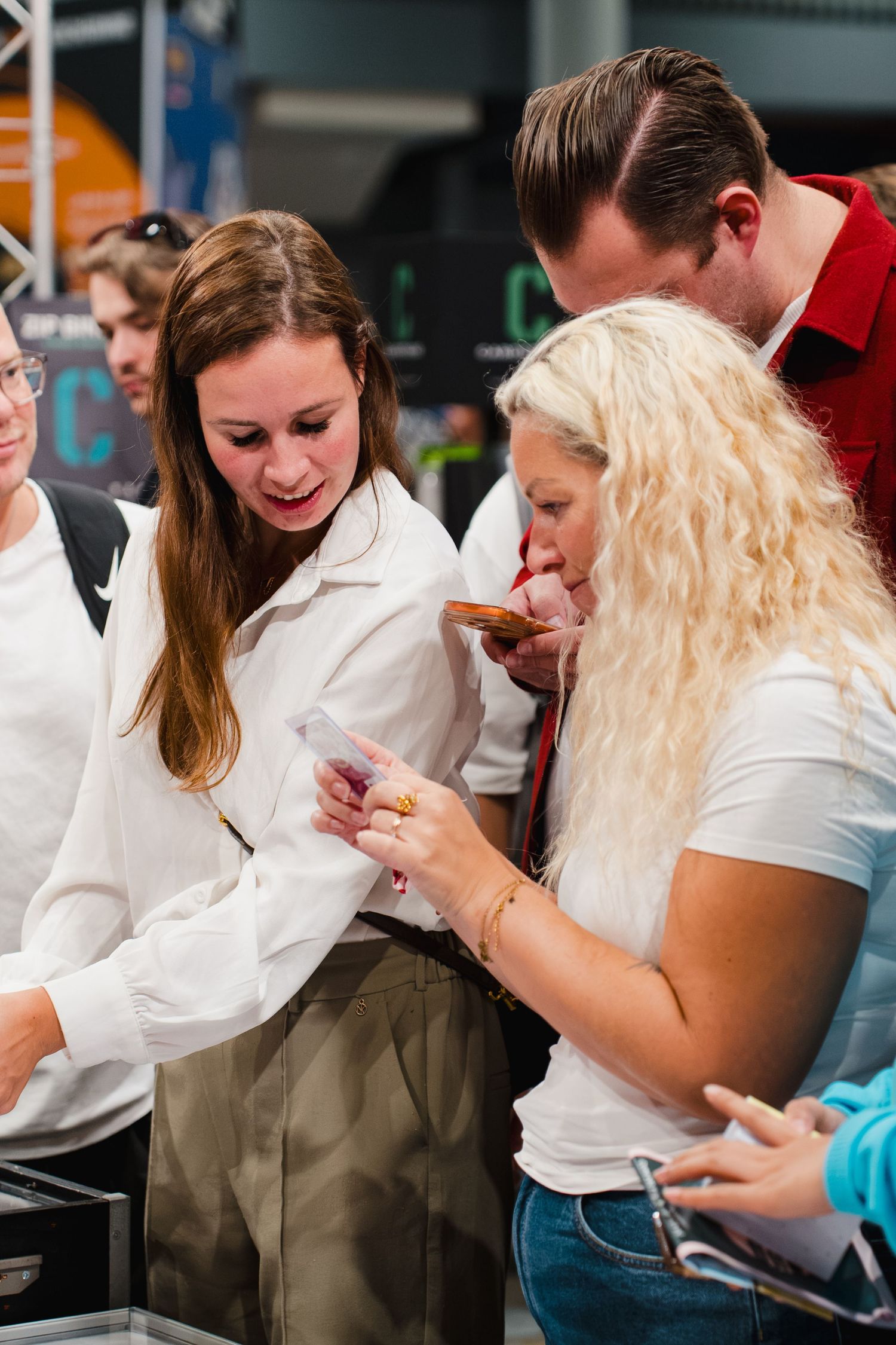 Two women examine a small food item at an indoor event, surrounded by other attendees in a bright venue.