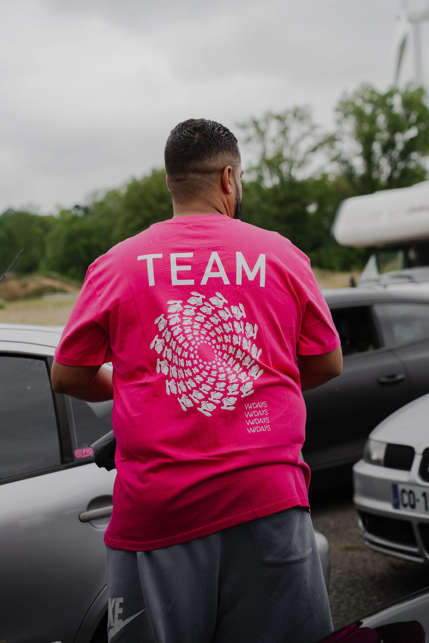 Man wearing a pink TEAM t-shirt standing near cars outdoors.