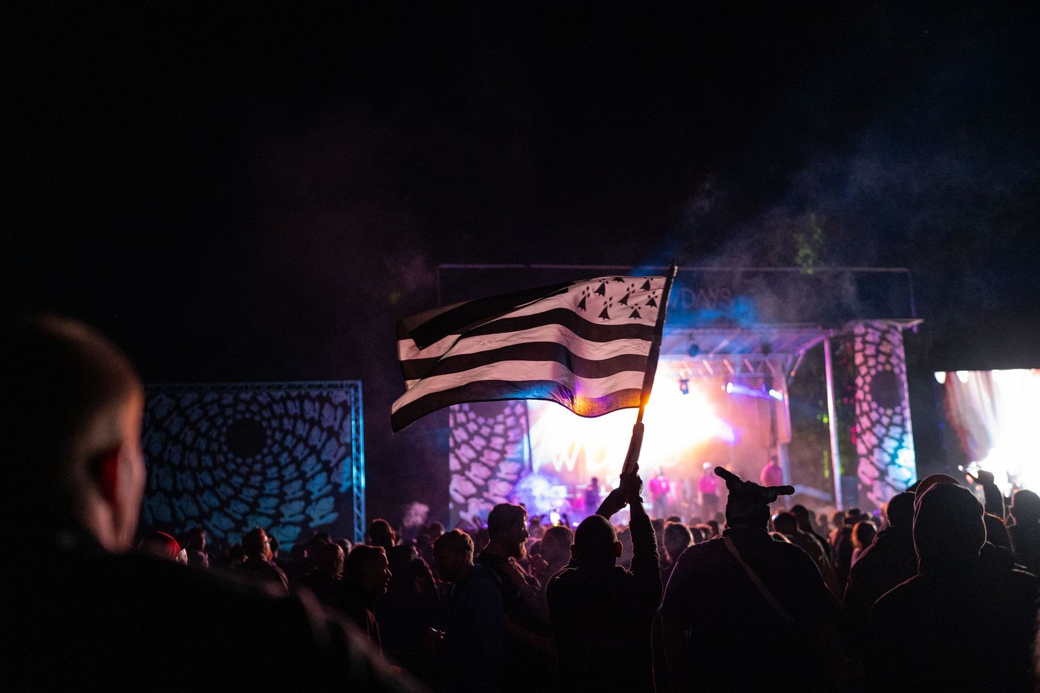 Crowd at outdoor music festival at night with person waving a Breton flag near illuminated stage.