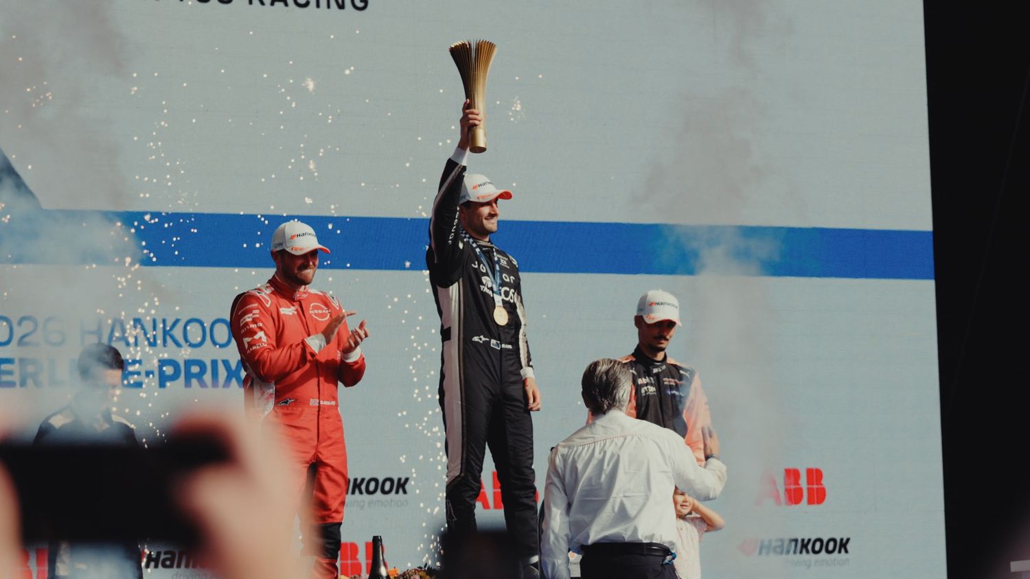 Formula E race winner raises trophy on podium while runners-up applaud amid confetti celebration.