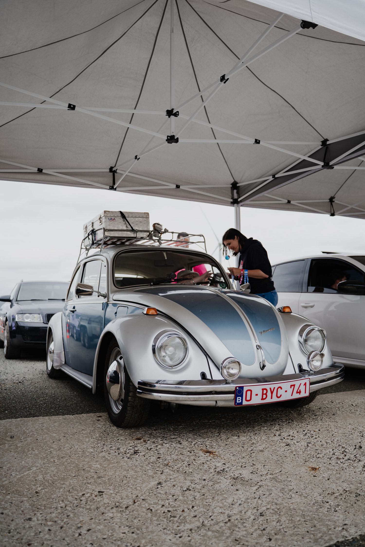 A classic silver VW Beetle with Belgian license plate parked at a car show under a white tent.