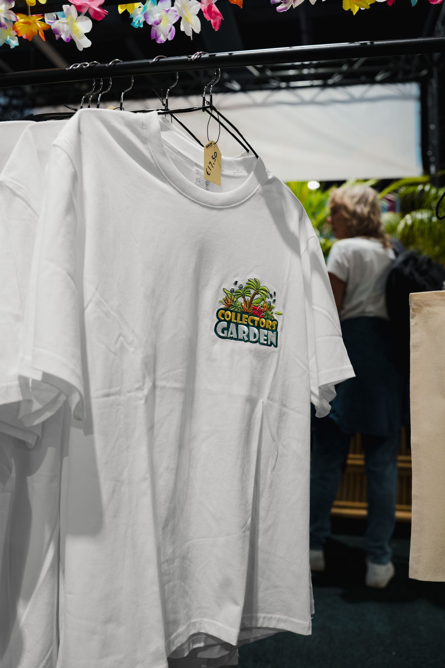 White t-shirt with Collectors Garden logo on chest, hanging on rack at outdoor market event.