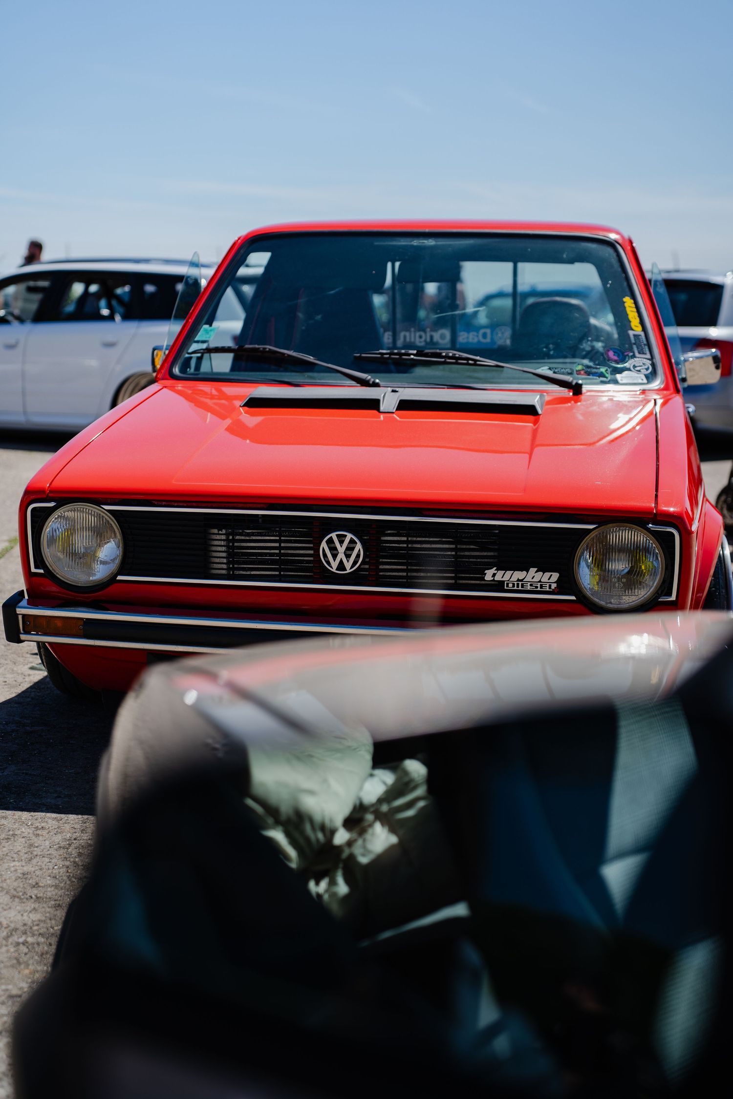 Red Volkswagen Golf Mk1 Turbo parked at a car meet under clear blue skies, front view showing VW badge.