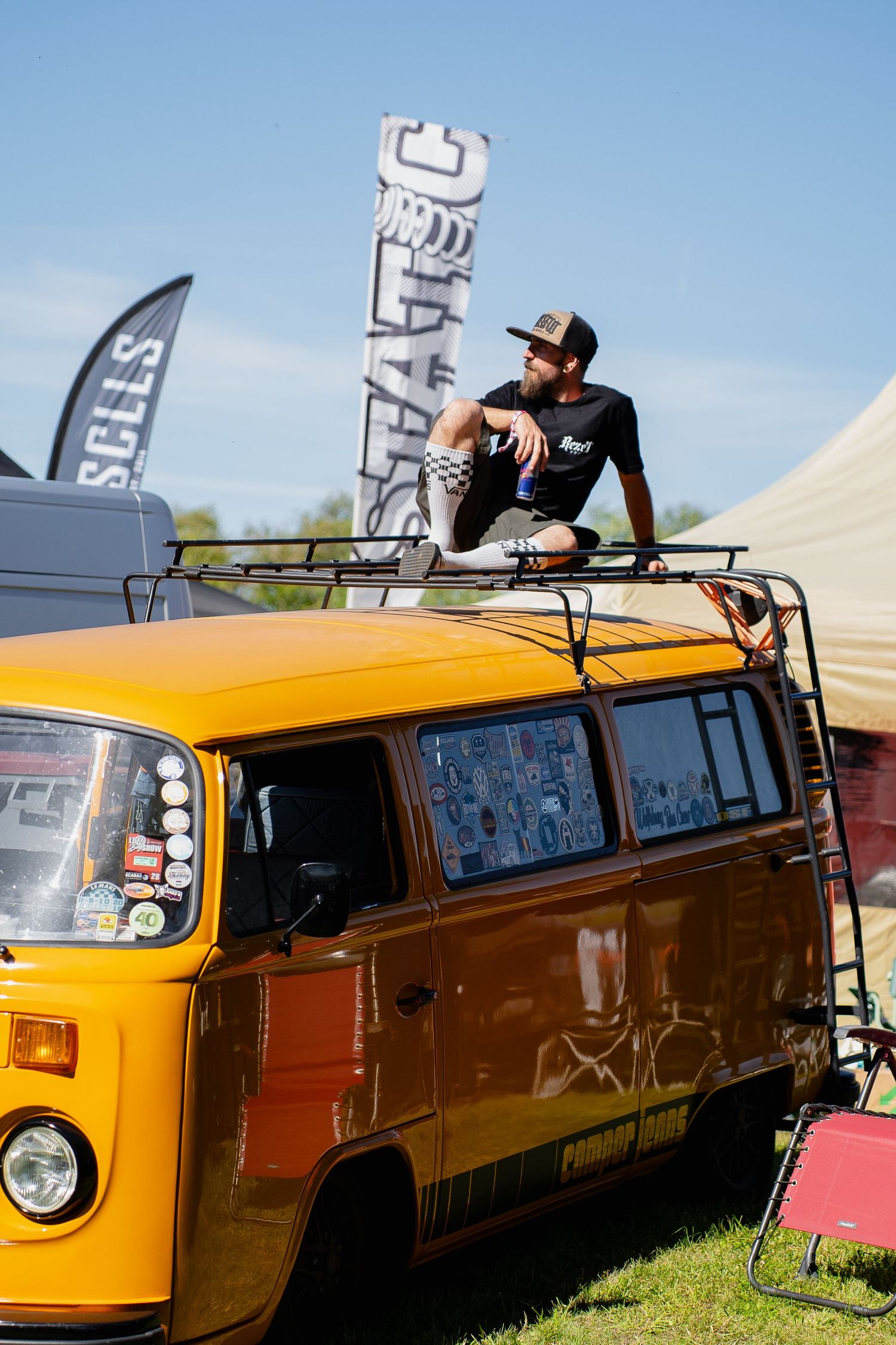 Man sitting on roof rack of vintage yellow Volkswagen van at outdoor car show on sunny day.