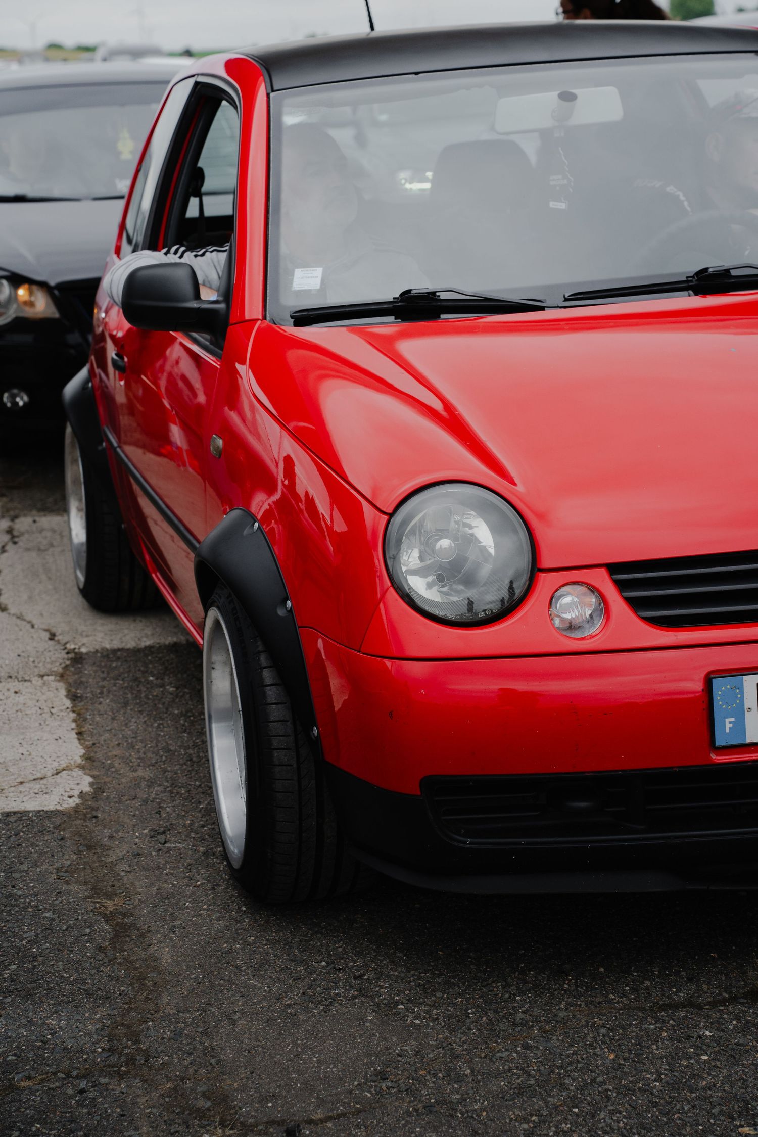 Red Volkswagen Lupo compact car parked on asphalt, featuring round headlights and custom wheels, with a black car visible behind.