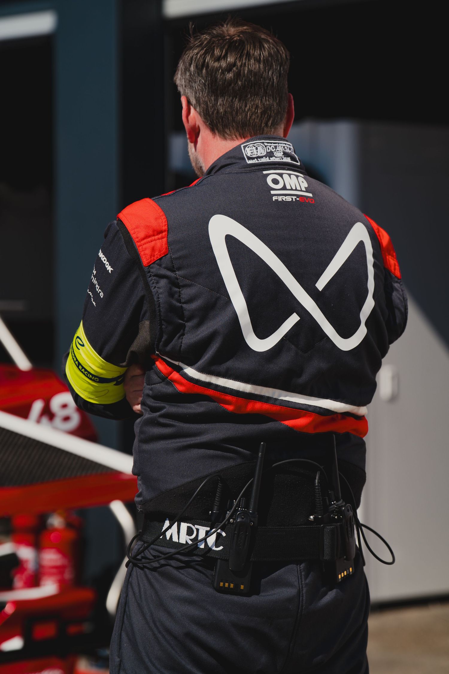 Racing team crew member wearing OMP black uniform with red accents and MRTC logo at motorsport event.
