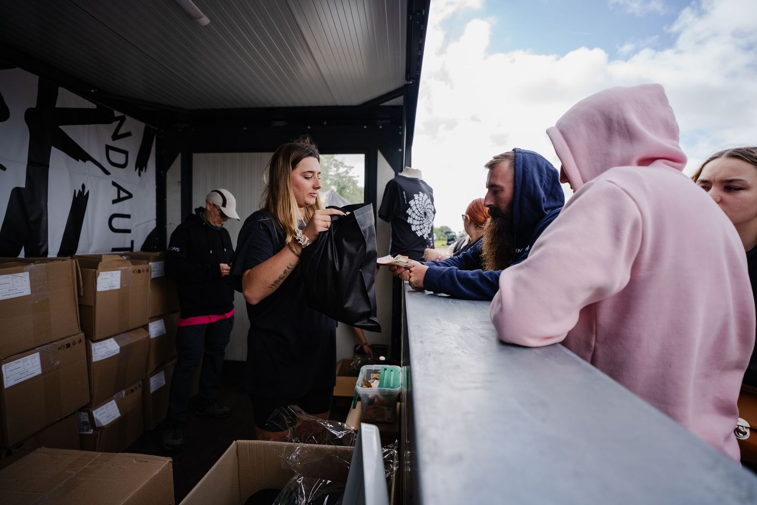 Woman selling merchandise from a vendor booth at an outdoor event to customers in hoodies.
