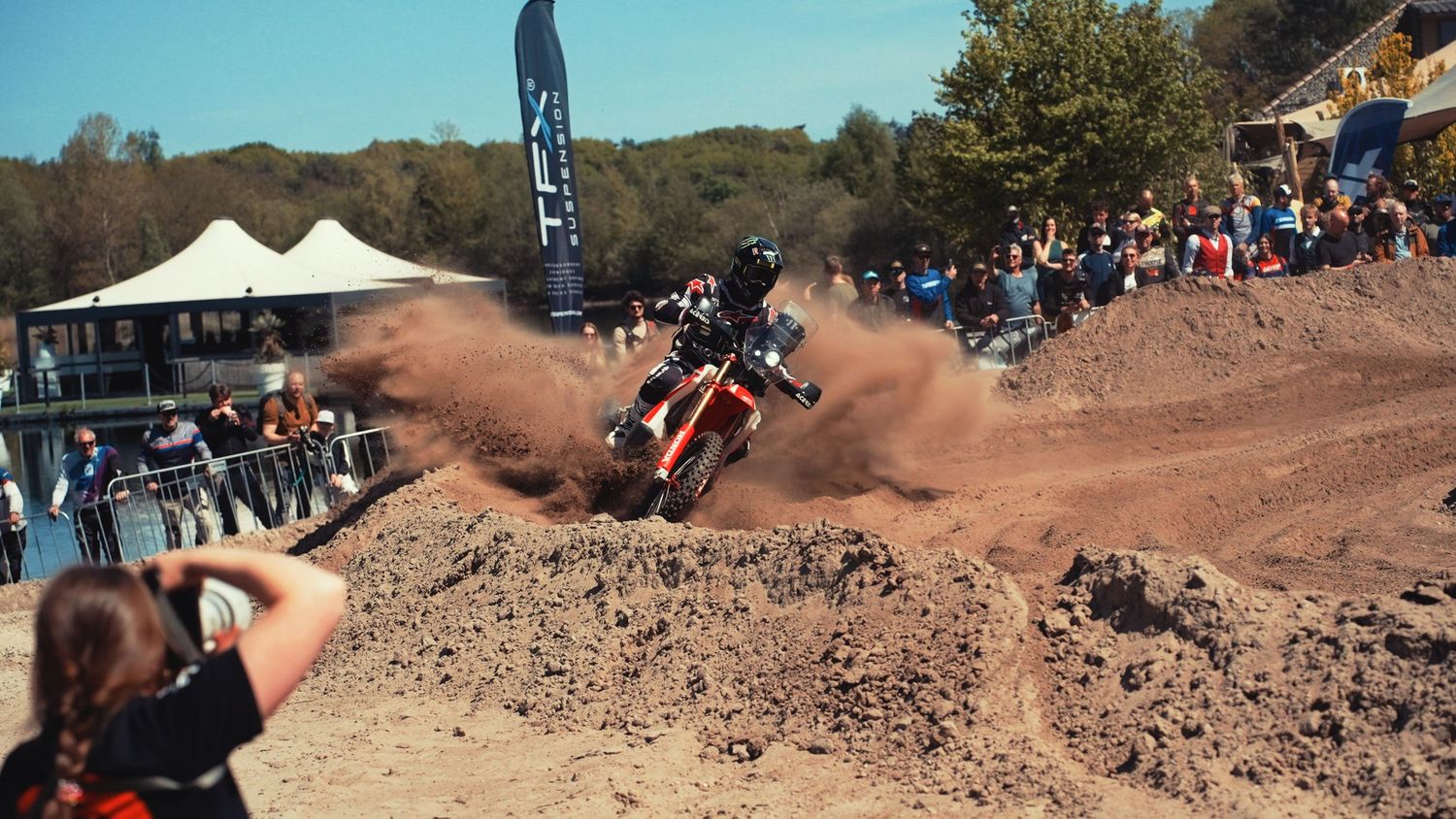 Motocross rider launching off a dirt jump at an outdoor competition event with spectators watching.