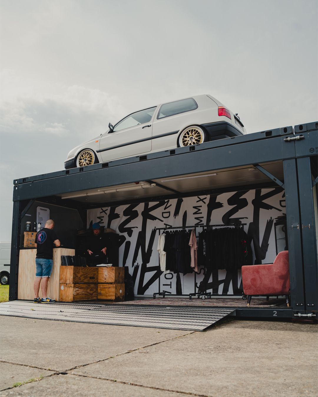 Modified white car displayed on top of black shipping container with graffiti artwork.