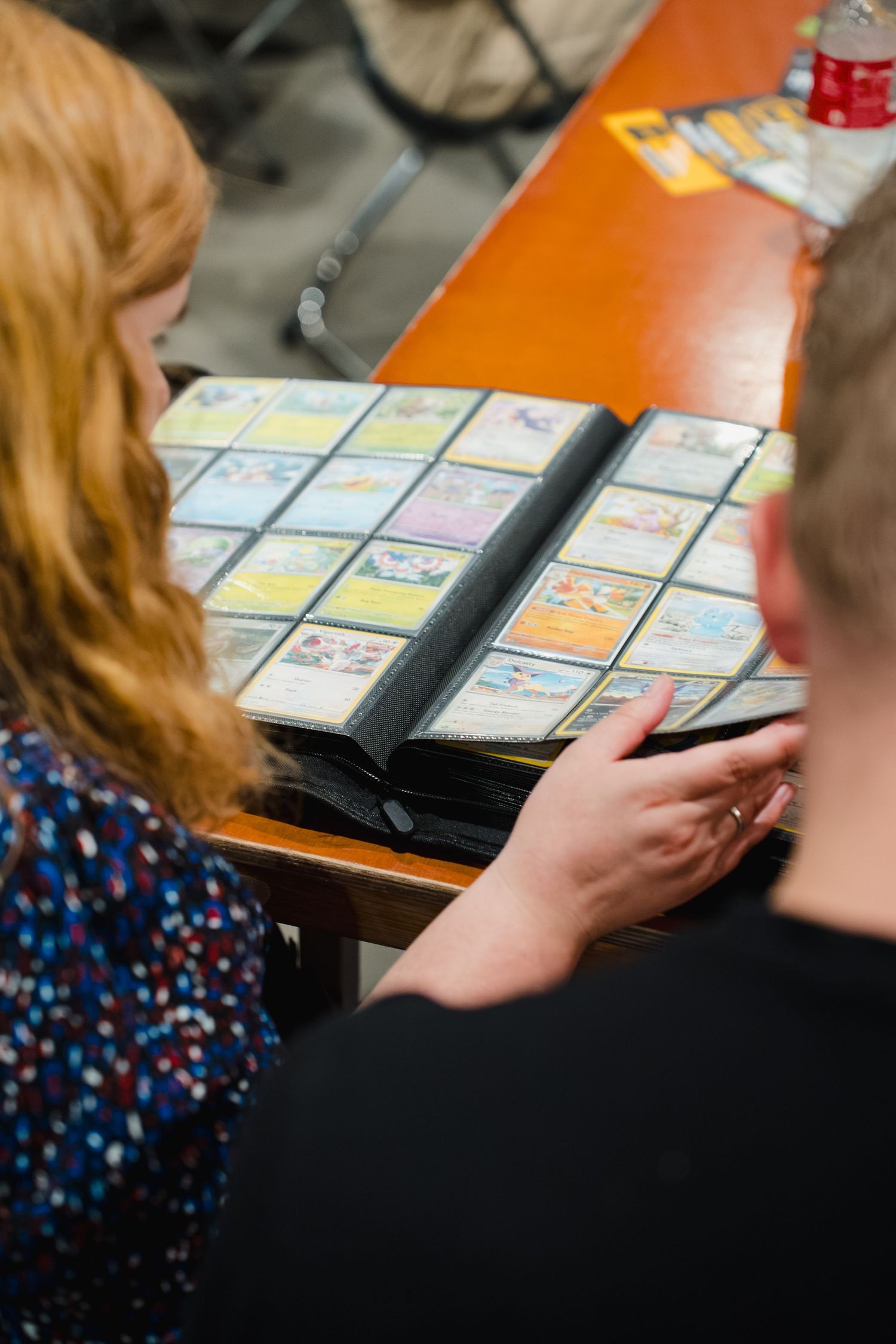 Two people browsing a Pokemon trading card collection album at a table during a card trading event.